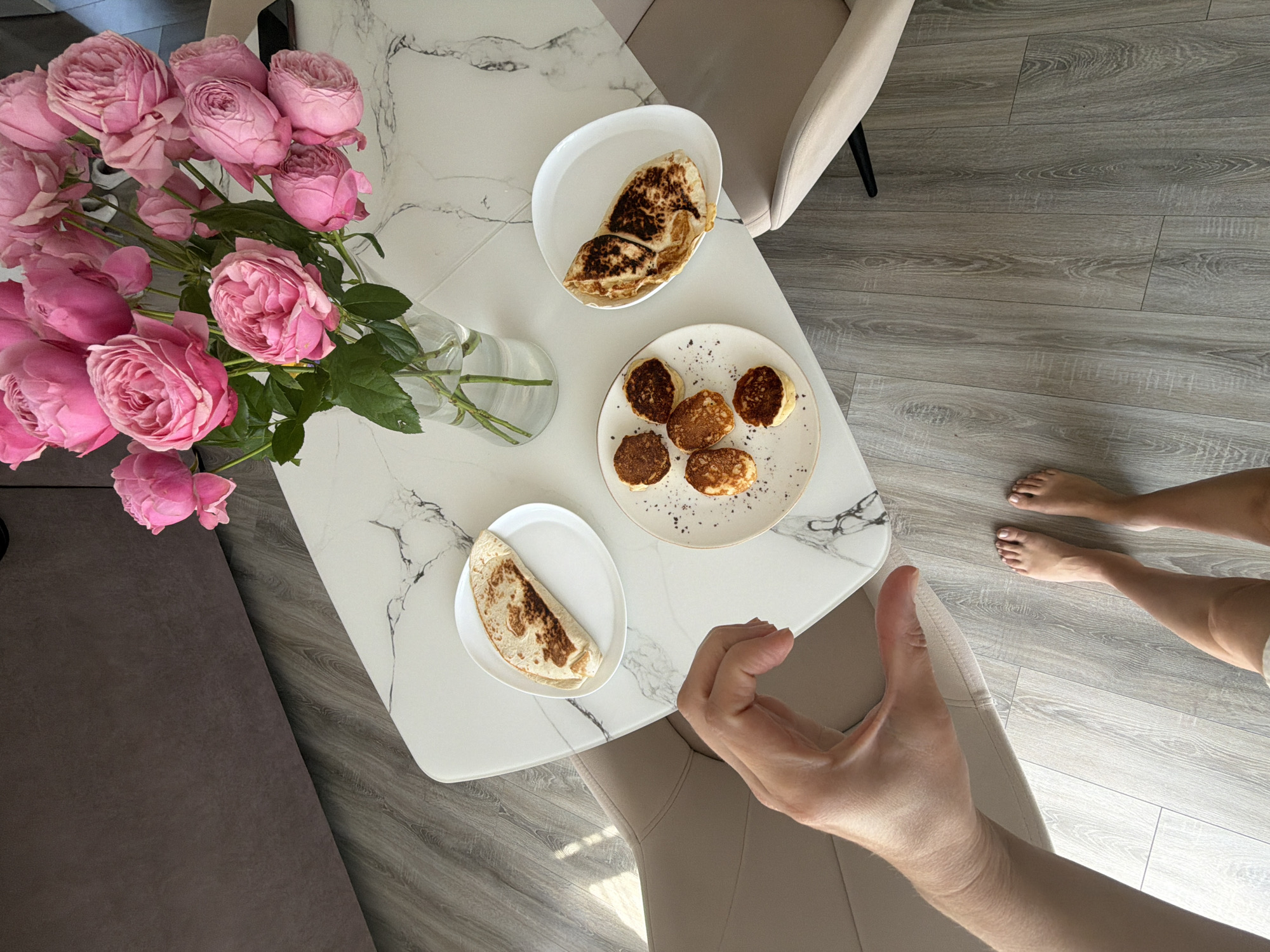 In Malinovka, Russia, a high-angle shot captures a marble-topped dining table laden with breakfast items. A bouquet of pink roses in a glass vase dominates the center. To the left, a white plate holds a folded quesadilla, while to the right, another white plate displays several small, golden-brown pancakes. In the foreground, a bare left foot and toes of a person are visible, standing on a light grey wood-planked floor. A left hand, positioned centrally, forms a heart shape with its thumb and index finger. Two upholstered chairs, one in the foreground and another in the background, flank the table. A dark grey countertop or cabinet is visible in the upper left corner.