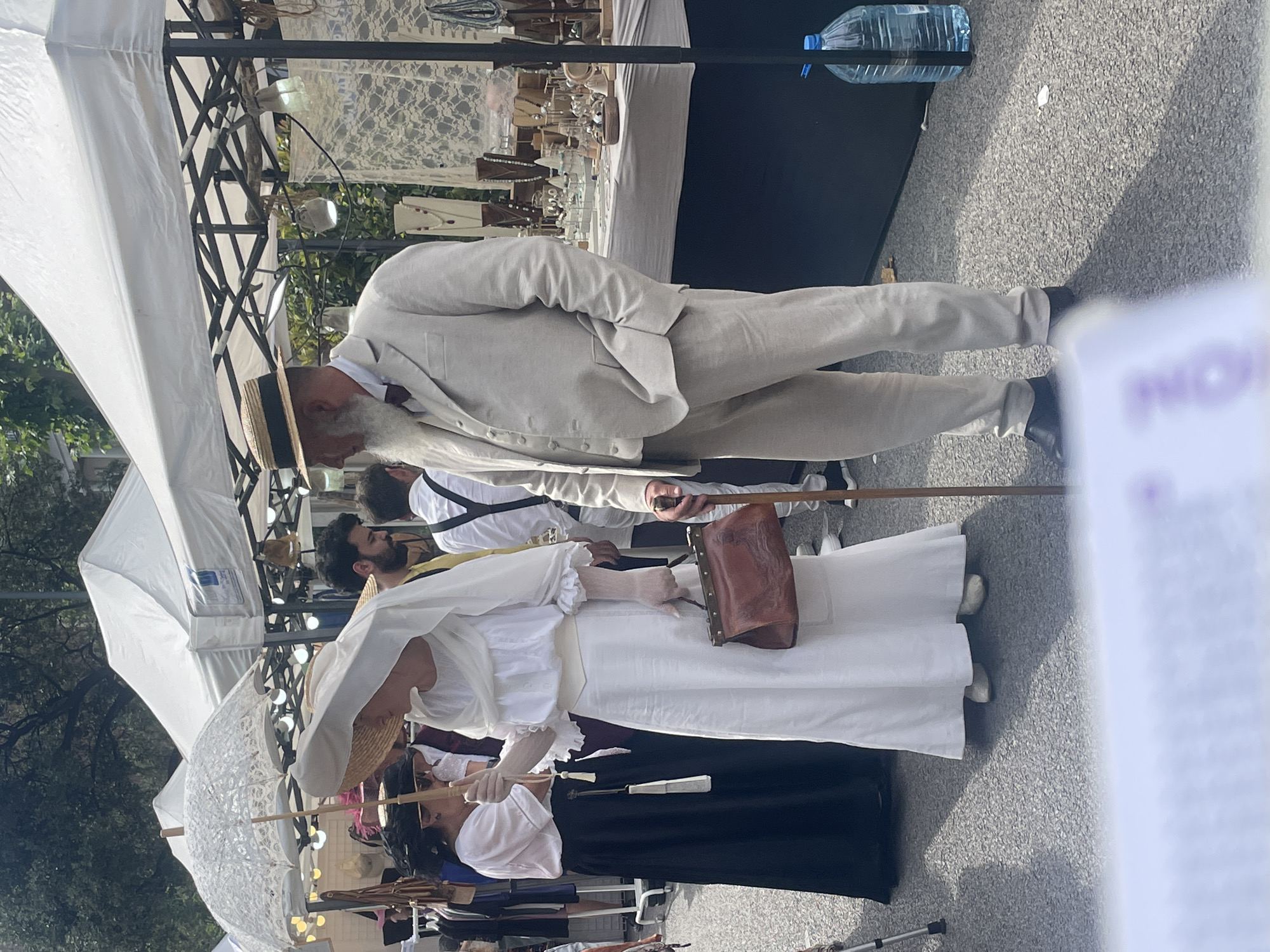 A daytime outdoor scene shows a group of people dressed in what appears to be historical clothing. The main subjects include a man in a light-colored suit and hat, with a walking stick, and a woman in a long white dress, black skirt, and hat, carrying a brown bag and a decorative white umbrella. They are standing among other people, some of whom are also in period attire. They are at what looks like an outdoor market. The background features market stalls with white canopies and displays of goods. The location is most likely in Barcelona, Spain.