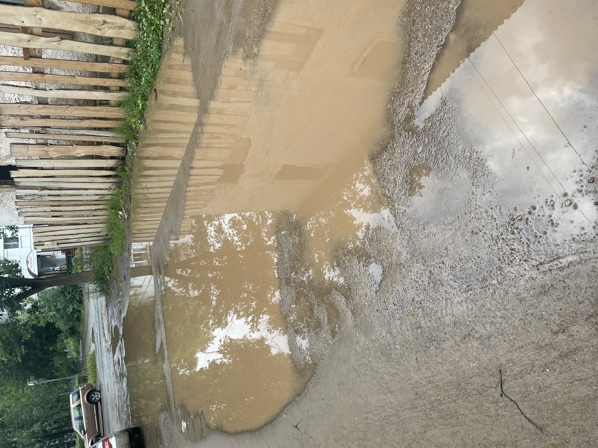 The image depicts a wet, possibly rain-soaked, outdoor scene. Dominating the foreground is a large puddle of muddy water reflecting the sky, trees, and what appear to be wooden planks or a fence. The ground is a mix of gravel and dirt, with some areas appearing more compacted and wet than others.
To the left, a wooden fence constructed from horizontal planks runs along the edge of the frame. Behind the fence, a white building with some signage is visible. Green grass and foliage are present along the base of the fence. A tree with dense green leaves is also visible in this area.
Further down the path, a light-colored car is parked. The overall impression is of an urban or semi-urban environment that has recently experienced rain. The sky in the reflection appears overcast, suggesting a cloudy day. The time of day is indeterminate but likely daytime given the ambient light. No people are visible in the image. There is some text visible on the white building, partially obscured by shadow and reflection, that appears to read "РЕМОНТ," which translates to "REPAIR" in Russian. The scene is located in Chaikovsky, Russia.