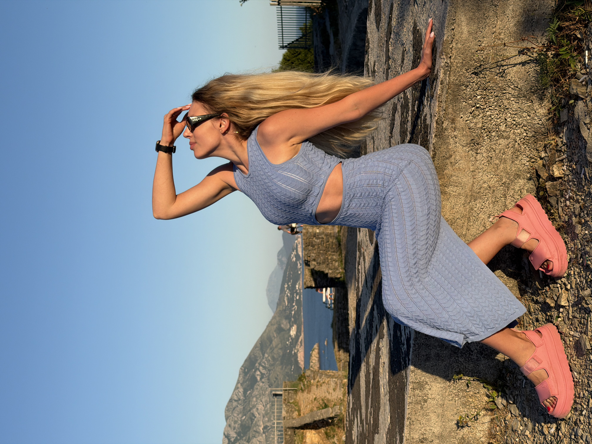 The image displays a lone female seated on a rough stone ledge at an elevated coastal viewpoint in Budva, Montenegro, under a clear, bright blue sky. The female has long, straight blonde hair and a tan complexion. She is attired in a light blue, two-piece knitted outfit, comprising a sleeveless crop top with midriff cut-outs and a matching midi-length skirt, both featuring a distinct chevron-patterned knit. She wears pink platform sandals with two wide straps. Her accessories include black-framed sunglasses bearing "VOGUE" on the temple, small gold-colored drop earrings, and a dark-colored smartwatch on her left wrist. Her fingernails are light-colored, and her toenails are painted red.
The woman is positioned in profile, her head tilted upwards and backward, gazing into the sky. Her left arm is raised, with her hand covering her forehead, as if shading her eyes. Her right arm is extended backward, resting on the textured stone surface for support. Her left leg is bent, and her right leg is extended.
The immediate foreground consists of the coarse, greyish-brown stone ledge and a sparse, stony ground cover with small patches of dry vegetation. Elongated shadows are cast from the left, indicative of a low sun angle. In the midground, additional ancient-looking stone structures and a metal railing are visible. Further back, blurred figures of other individuals are discernible, including one seated on a bench. The background reveals a vast, unobstructed blue sky above the Adriatic Sea. Rugged mountains emerge directly from the sea, their lower slopes adorned with scattered white-roofed buildings typical of coastal settlements along the Montenegrin Riviera, confirming the location as Budva.