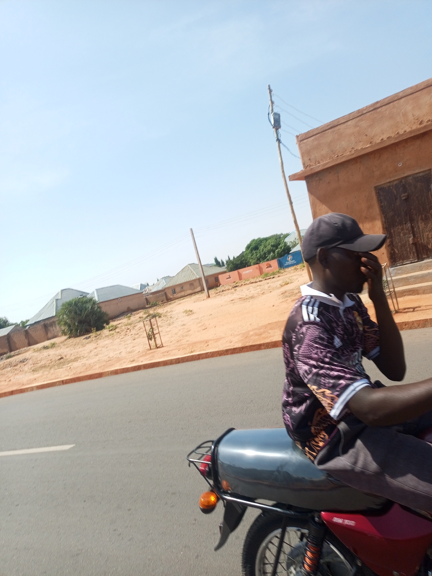 A man wearing a baseball cap and patterned short-sleeved shirt is riding a red motorcycle on a paved road. He has his hand to his face. The road has a median on one side with dry, sandy ground. In the background are single-story buildings, and a dirt yard. There is an electrical pole in the distance. The sky is blue and clear.
