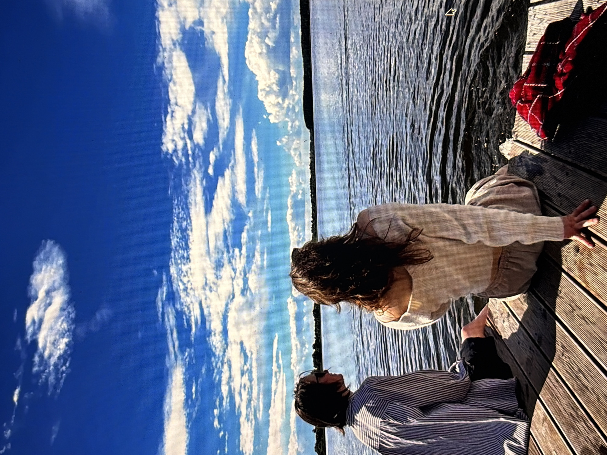 The image depicts two individuals, viewed from behind, seated on a wooden pier. They are looking out at a vast body of water under a bright blue sky dotted with white, puffy clouds. The person on the left wears a vertically striped long-sleeved shirt and dark shorts, with one foot extended towards the water. The person on the right is dressed in a light-colored, off-the-shoulder sweater and light-colored pants. A red and black plaid blanket is folded on the pier to the right of the second person. The water exhibits gentle ripples, and a distant tree line forms the horizon. The scene is bathed in sunlight, casting shadows on the wooden planks of the pier. The location is not identifiable as Moscow, Russia, based on the visual information provided.