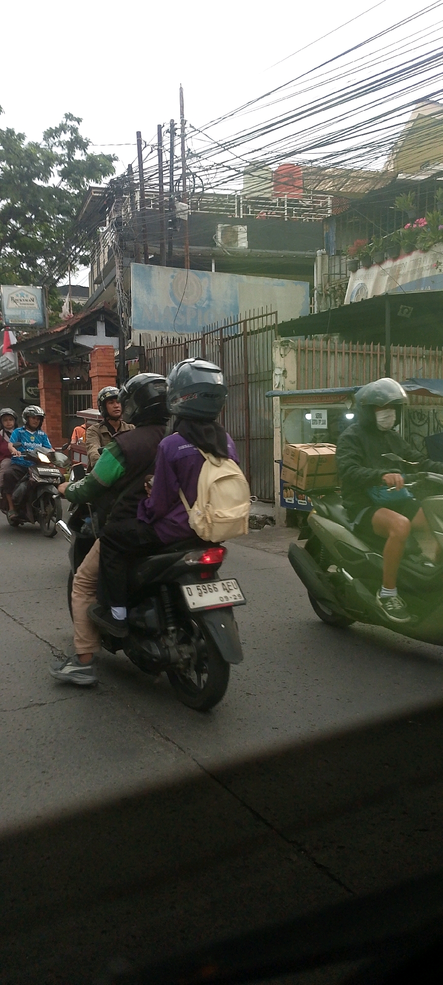In Bandung City, Indonesia, on a paved road with visible cracks and some debris, a group of motorcyclists is traveling. The foremost motorcyclist is wearing a purple jacket and a beige backpack, with a dark helmet. Behind them, another motorcyclist is partially visible, wearing a dark jacket and helmet. Further ahead, on a parallel path or lane, a motorcyclist in a dark jacket and helmet is riding a lighter-colored scooter. To the left, three individuals are on a motorcycle; the driver wears a helmet, and the two passengers, one in a blue jersey and another in a helmet, are also visible. A red and white Indonesian flag is mounted on a pole near a building to the left. In the background, there are buildings with metal fences, overhead electrical wires, and trees. A vendor cart selling boxed goods is positioned to the right of the road. The overall scene depicts urban street life with a focus on motorcycle transportation.