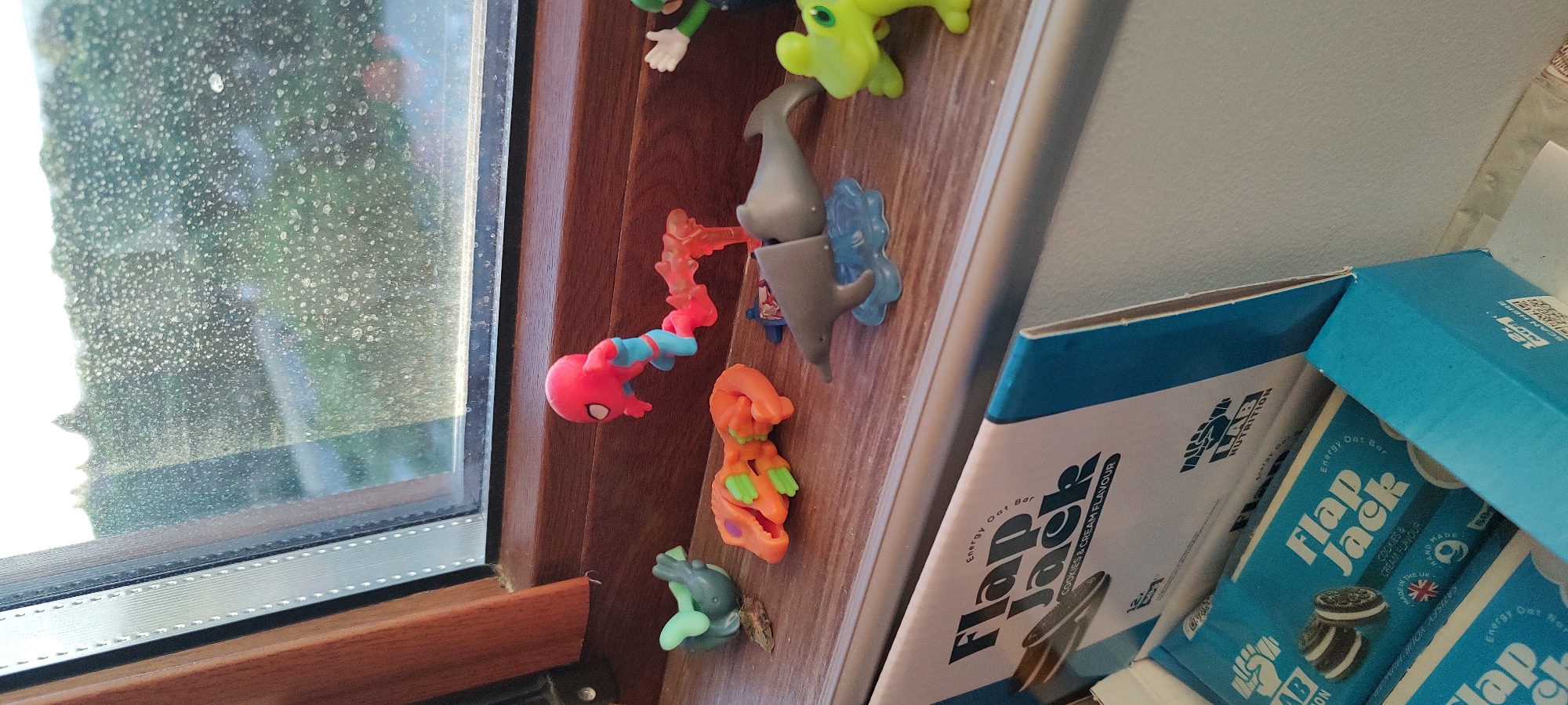 The image shows a variety of objects. A window, possibly in a domestic setting, appears on the left, the glass obscured with water stains or condensation. To the right of the window frame, there are several small plastic toys mounted on a wooden surface. These figures include what looks like a Spider-Man toy, a shark-like figure, and other colorful, cartoonish toys. Next to the toys, there are several boxes of "Flapjack" energy bars stacked together. The brand name is visible on the packaging, indicating they are oat bars with a "Cookies & Cream Flavor." The image is closely framed, providing a detailed view of the objects and textures.