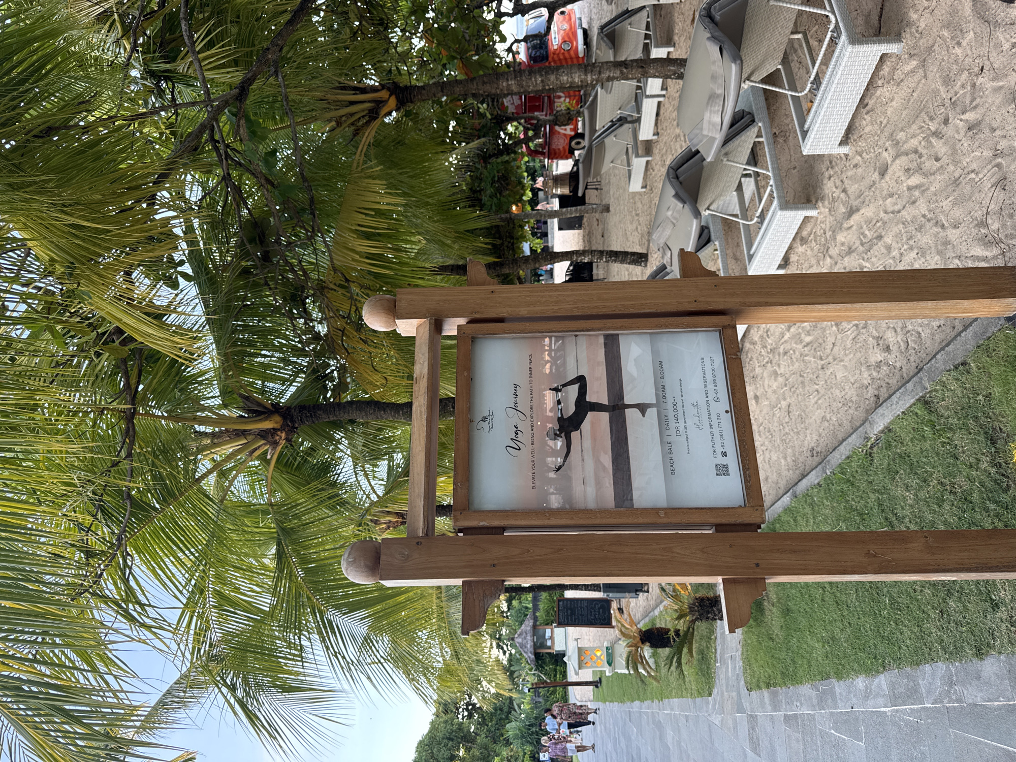 This outdoor shot features a wooden sign advertising a "Yoga Journey" held daily at the Beach Bale from 7:00 AM to 8:00 AM, costing IDR 140,000++. The background shows a beach setting with lounge chairs and palm trees. A stone pathway stretches behind the sign, lined with grass, more palm trees, and glimpses of structures in the distance. People are walking on the pathway. The location is an unidentified area within the city of Unknown, Unknown.