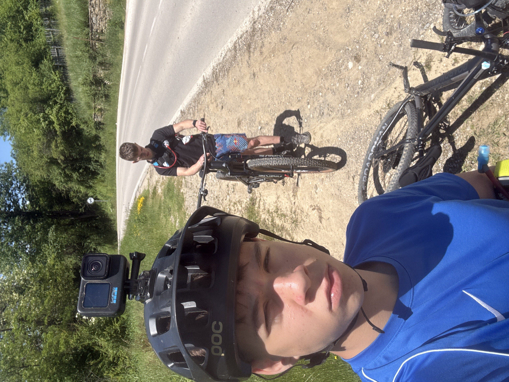 The image shows two male cyclists pausing on the side of a paved road. The foreground subject is taking a selfie while wearing a blue jersey, a black POC helmet with a GoPro camera mounted on top. The road is adjacent to a grassy area with trees. In the background, another cyclist is standing with his bike and looking at a device on his wrist. They are presumably in the area of Donchovci, Bulgaria, although specific landmarks are not visible to confirm the exact location. The scene suggests a break during a cycling ride.