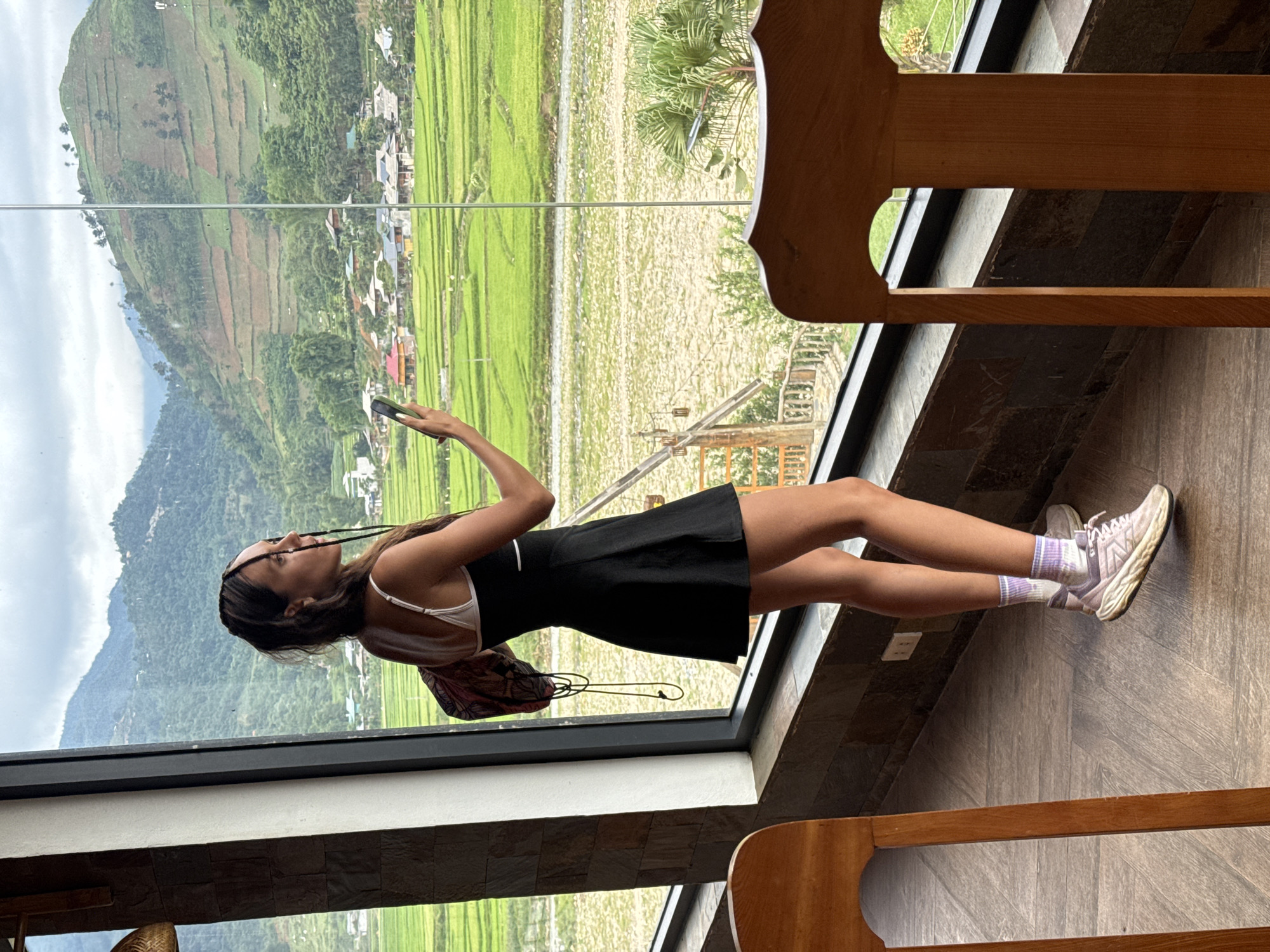 A woman stands in front of a large window, taking a picture with her phone of the view in Tú Lệ, Vietnam. The window overlooks green rice fields, a road, and mountains. The woman is wearing a black dress, white tank top, and sneakers. She is holding her phone up to take a picture. She has a black cord hanging down her chest. The background shows a landscape of rice terraces, houses, and mountains under a cloudy sky. The scene is indoors with wooden floors and chairs.