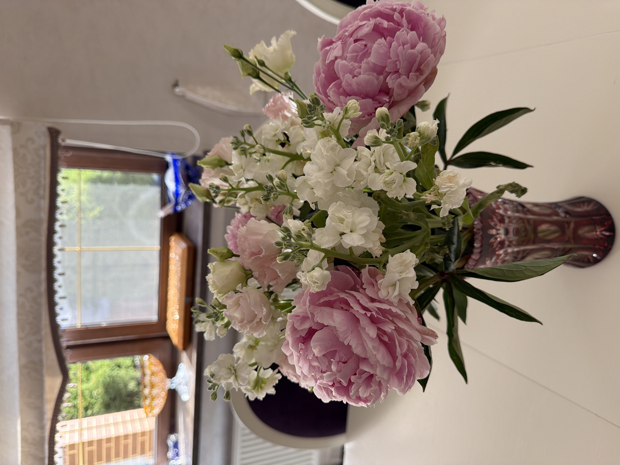 The image depicts a floral arrangement of pink peonies and white stock flowers in a red glass vase, placed on a white table. In the background, a window reveals greenery and trees, suggesting an outdoor setting. The window has a wooden frame and a curtain. Also visible are a decorative dish with a yellow, translucent substance and a blue ceramic object. The interior appears to be a dining area, with a portion of a chair visible to the left. The overall scene is one of domestic tranquility. The image is not in the city of Mishutkino, Russia.