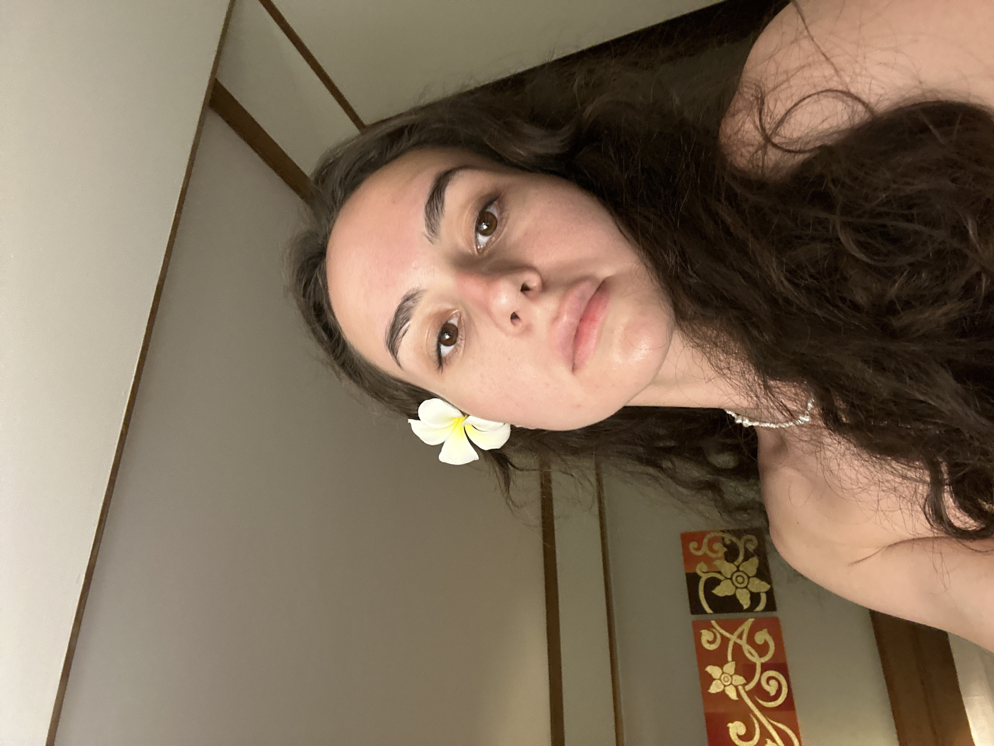A person with long, dark brown hair is facing the camera, with a white plumeria flower behind their left ear. They have fair skin, brown eyes, and slightly pursed lips. They appear to be wearing a pearl necklace. The background is a wall, partially showing two decorative wall hangings with a gold floral design on a dark brown and red background, respectively. The location is inferred to be Pattaya City, Thailand, due to the presence of the plumeria flower which is often associated with tropical regions like Thailand.