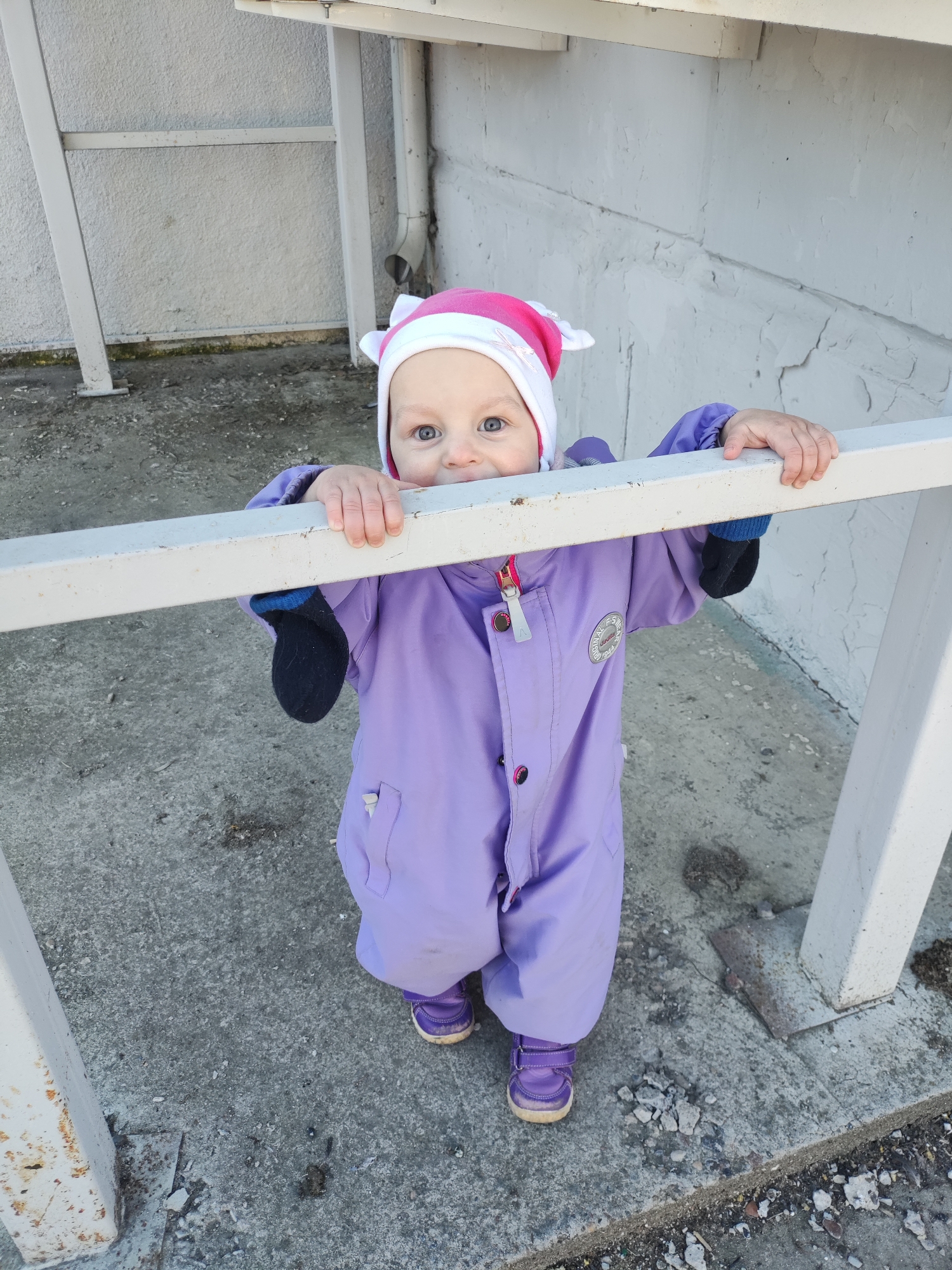 A young child, appearing to be a toddler, is captured in a full-body outdoor shot, looking directly at the camera with wide blue eyes. They are dressed in a bright, light purple one-piece jumpsuit with a visible zipper and snap buttons, and a circular patch on the left side of the chest (text on patch is illegible). On their head is a pink and white hat with ear-like features. The child wears purple shoes.
The child is standing behind and holding onto a horizontal white metal railing with both hands, peeking over it. The railing shows some signs of wear and rust. The background consists of a textured, light-colored concrete wall, with a grey drainpipe visible on the right side. The ground is also concrete, appearing cracked and somewhat dirty with small debris.
The image is taken in bright, natural daylight, suggesting it is daytime. The child has a curious and engaging expression. The setting appears to be an urban or semi-urban outdoor area, possibly a walkway or entrance to a building in Chaikovsky, Russia.