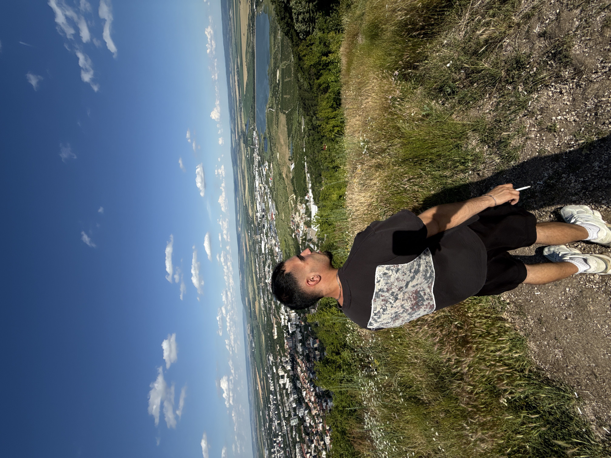A man is sitting on a grassy hillside overlooking a city. He is wearing a black t-shirt with a patterned design on the front, black shorts, and white sneakers. He is holding a cigarette in his right hand. The sun is shining and the sky is blue with scattered clouds. The city below is spread out, with buildings and roads visible. There are trees and greenery on the hillside and in the distance. The overall impression is a relaxed and scenic moment. The image appears to be taken in Shumen, Bulgaria, on a clear day.