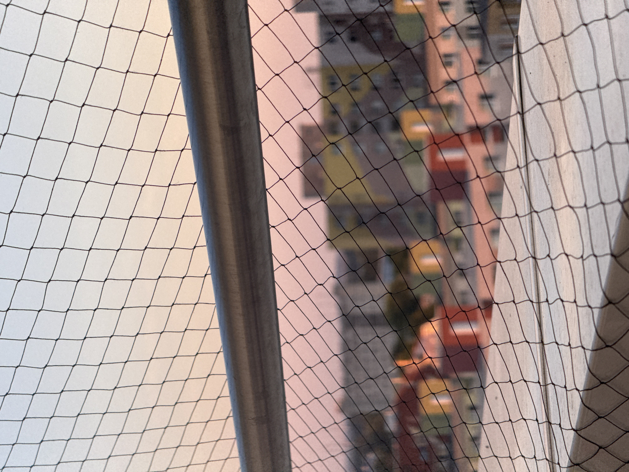 The image shows a view through a black safety net. In the foreground, a grey metallic rail runs horizontally across the image, with the net draped over it. Below the rail, the net obscures a blurry expanse of colorful buildings, suggesting an urban setting. The sky above the buildings is a gradient of soft pink and light blue, indicative of either sunrise or sunset. There are no people or animals visible in the image. The scene appears to be from a balcony or elevated vantage point, looking out over a cityscape at dusk or dawn. The specific location of Bratislava, Slovakia, is not discernible from the visual information presented in the image.