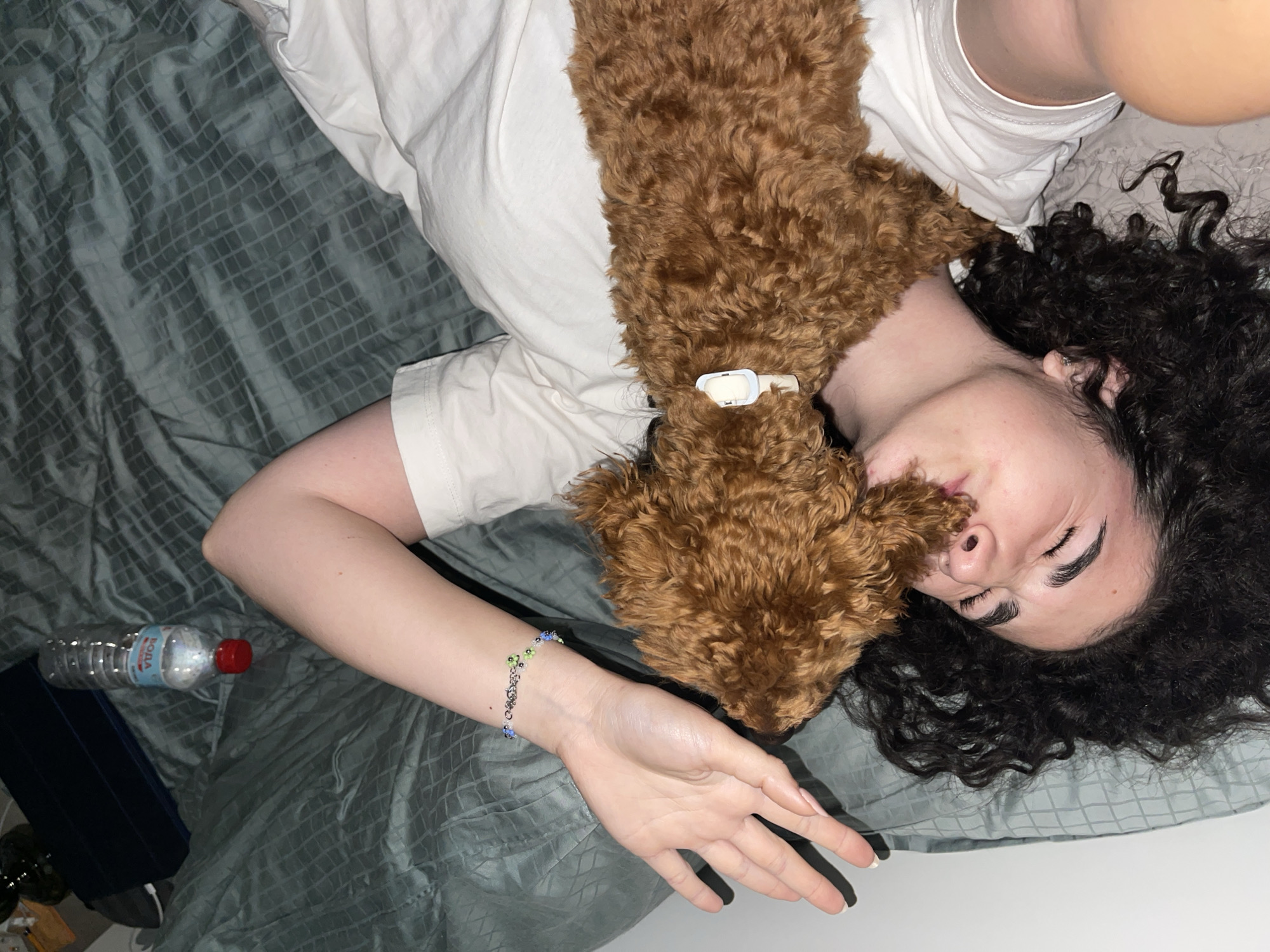 The image captures a young woman and a small, brown, curly-haired dog lying together on a bed in what appears to be an indoor setting, likely a bedroom in Sofia, Bulgaria. The woman is lying on her back with her head on a dark green or teal patterned pillow, her dark, curly hair fanned out. She is wearing a plain white t-shirt, and a colorful beaded bracelet is visible on her left wrist. Her eyes are closed or partially closed, suggesting a state of relaxation or sleep.
The dog, which has fluffy brown fur and wears a light-colored collar, is nestled affectionately on her chest and neck, its head close to her face. This intimate pose conveys a strong bond and comfort between the two subjects.
The bedding is a distinct dark green/teal with a subtle quilted or checkered pattern. Other objects visible include a clear plastic water bottle with a red cap, labeled "BORA," and a dark-colored tablet or device case to the left of the woman. The lighting is soft and ambient, contributing to a calm and peaceful atmosphere. The scene portrays a quiet moment of companionship and rest.