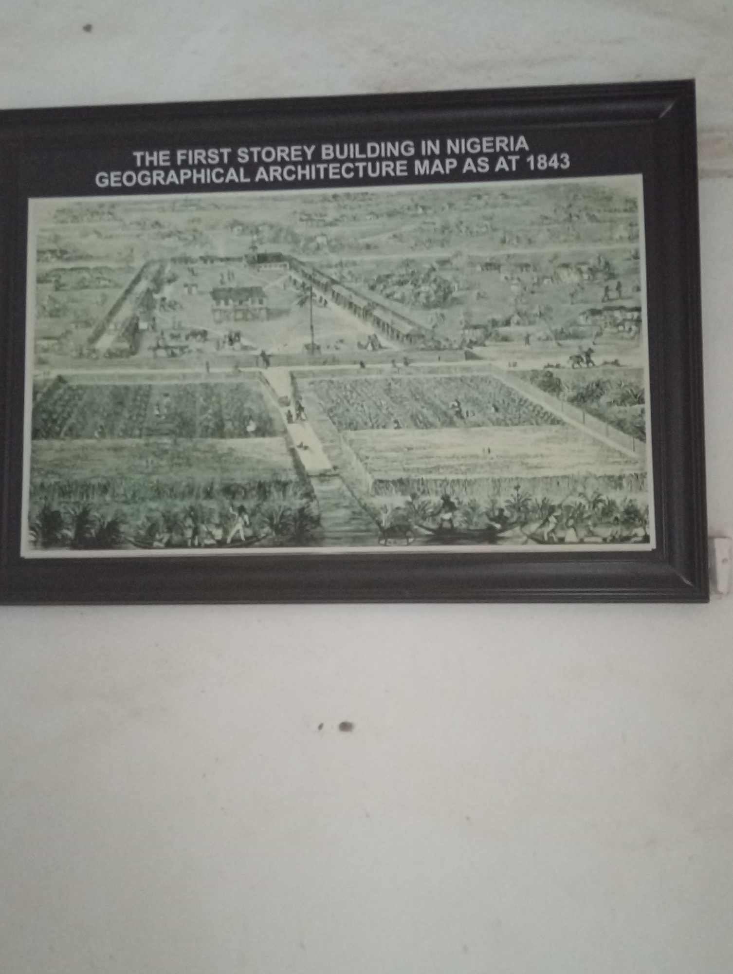 The image shows a framed print hanging on a white wall. The print depicts the "First Storey Building in Nigeria, Geographical Architecture Map as at 1843". It appears to be an early illustration or map showcasing the structure and surrounding area. The drawing shows a rectangular building with an open courtyard and several surrounding outbuildings. There are large cultivated fields with people working, pathways, and rows of crops that surround the structure. There are also people walking on the roads, and a few people can be seen in boats. The foreground shows detailed rows of vegetation.
It is located in the city of Badagry, Nigeria, representing a historical location.