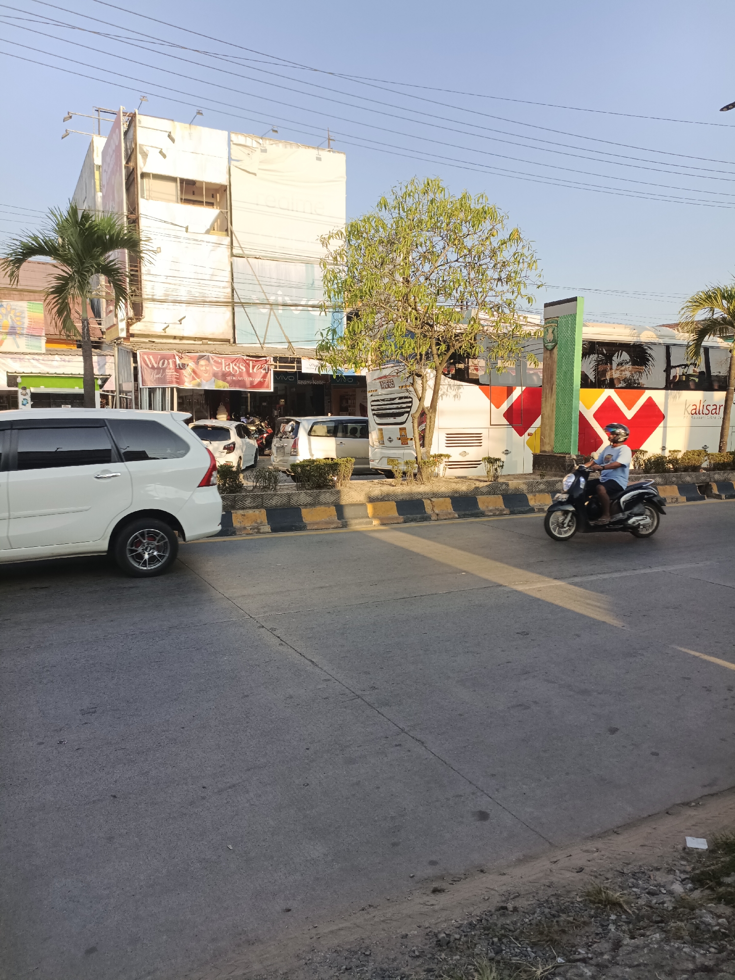 The image depicts a street scene in Sangatta, Indonesia, during the daytime. In the foreground, a white minivan is parked on the left side of the road, with its rear tire visible. A person wearing a blue shirt and a helmet is riding a black scooter in the right lane of the road, heading towards the viewer. In the background, a white bus with red and yellow heart-shaped graphics is parked. Several buildings line the street, including a white multi-story building with signs for "kalisar" and what appears to be a store. Palm trees and other foliage are also present. Power lines crisscross the sky above. The road is paved and appears to be in good condition, with yellow and black striped barriers separating the lanes. The overall atmosphere suggests a typical urban or commercial area.
