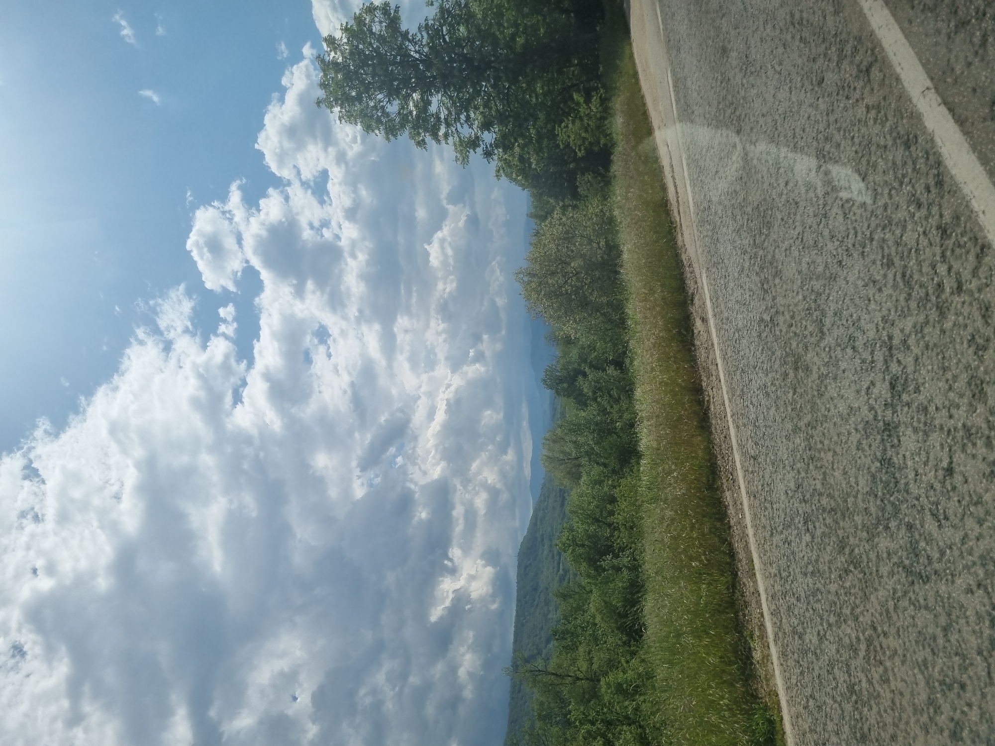The image depicts a view from a roadside. The sky is largely filled with white, billowing clouds against a blue background, with bright sunlight in the upper left corner. A line of green trees and vegetation separates the sky from the gray asphalt road on the right side of the frame. The road surface appears worn. In the distance, a hint of mountains is visible, suggesting an elevated or rural location. The image captures a natural landscape along a roadway. The location is likely Хаджийско, Bulgaria.