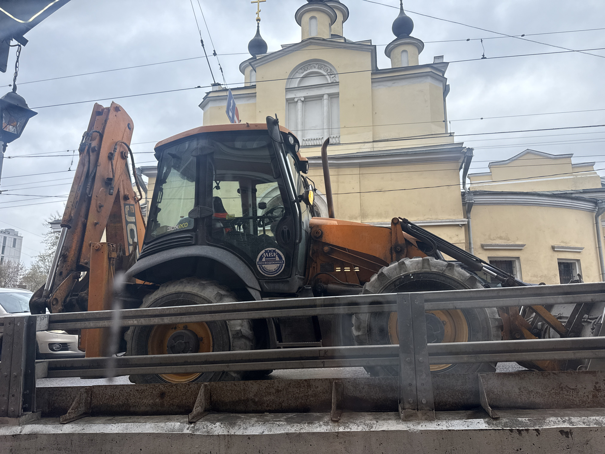 A backhoe is parked on what appears to be a trailer, with a pale yellow building in the background topped with dark domes and a cross, likely an Orthodox church. The backhoe is predominantly orange and black. A sticker is visible on the door of the cab with Cyrillic text, likely indicating a company name or service. Wires run overhead, suggesting an urban setting. The scene is overcast. The location is identified as Moscow, Russia by the presence of Cyrillic text and the distinctive Orthodox church architecture. A small flag, partially obscured, is attached to the top of the backhoe cab. A metal railing is positioned between the backhoe and the viewer.
