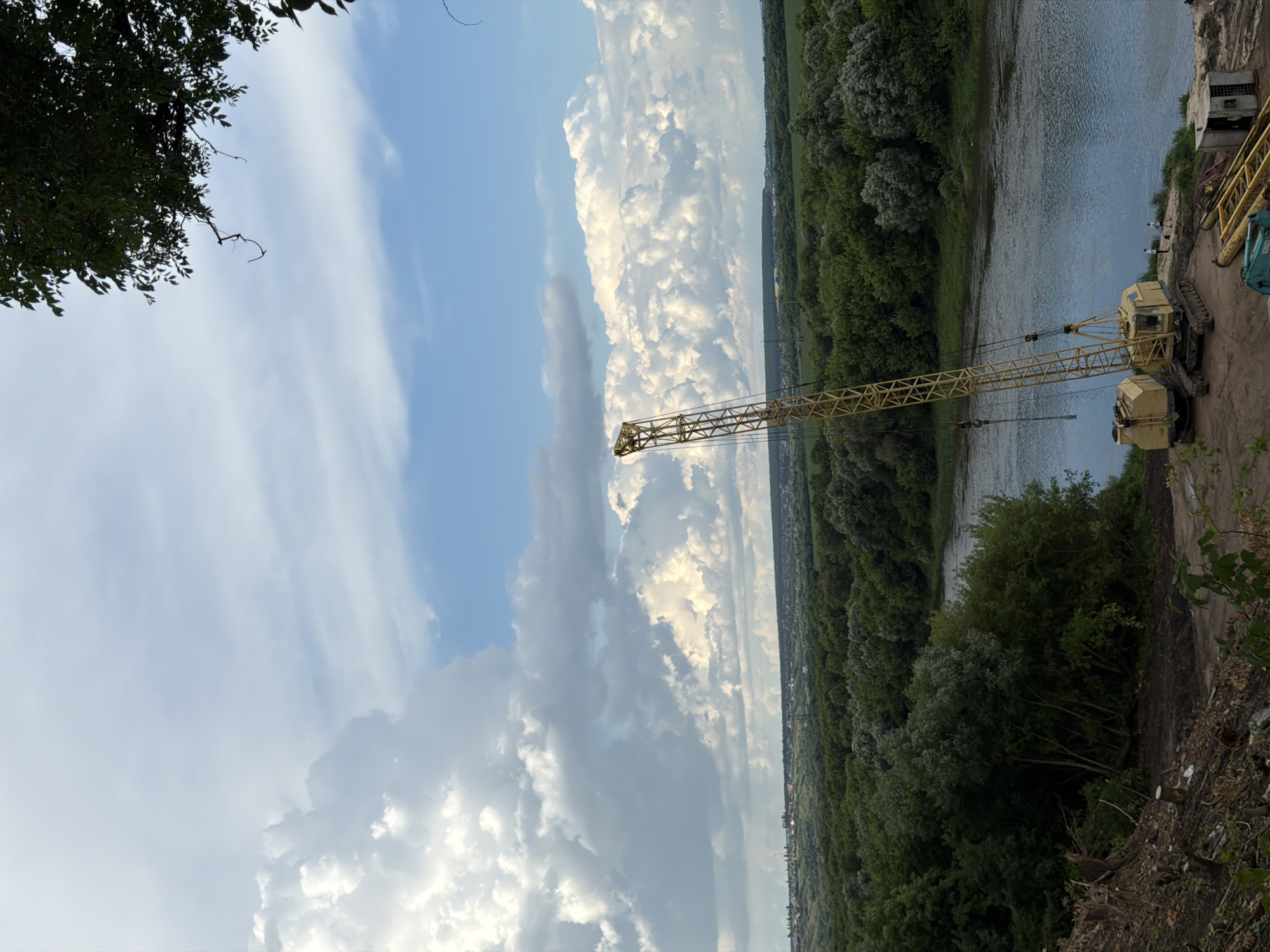 This is an outdoor, full-shot photograph taken from a high vantage point overlooking a river, a construction site, and a distant town. The sky is partially cloudy, with large white clouds against a blue sky. In the foreground on the left, there is a cleared area with tree stumps and debris, suggesting recent deforestation or construction preparation. Adjacent to this cleared area, a yellow crane with caterpillar tracks is positioned on the ground. Beside the crane, there are two larger yellow machinery units, one with a raised cabin and the other seemingly a counterweight or additional module. Further to the right along the riverbank, two individuals are standing near the water. The river itself is a calm, blue body of water. Across the river, a dense forest stretches into the distance, forming a green, rolling landscape. Beyond the forest, a town with buildings and structures is visible on a gentle hill under the horizon. The overall impression is a transitional scene, with signs of industrial activity set against a natural, pastoral backdrop. This scene is located in the city of Semiluki, Russia.