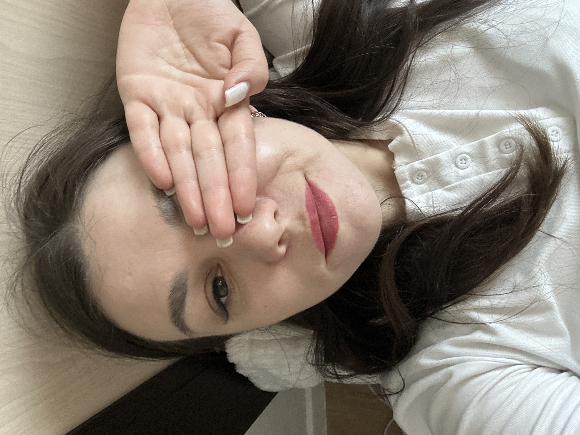 A young woman with long, dark hair is lying down, partially covering her face with her hand. She is wearing a white, button-down shirt. The background appears to be a light-colored wooden surface. The location is not identifiable as Chaikovsky, Russia from the given image.