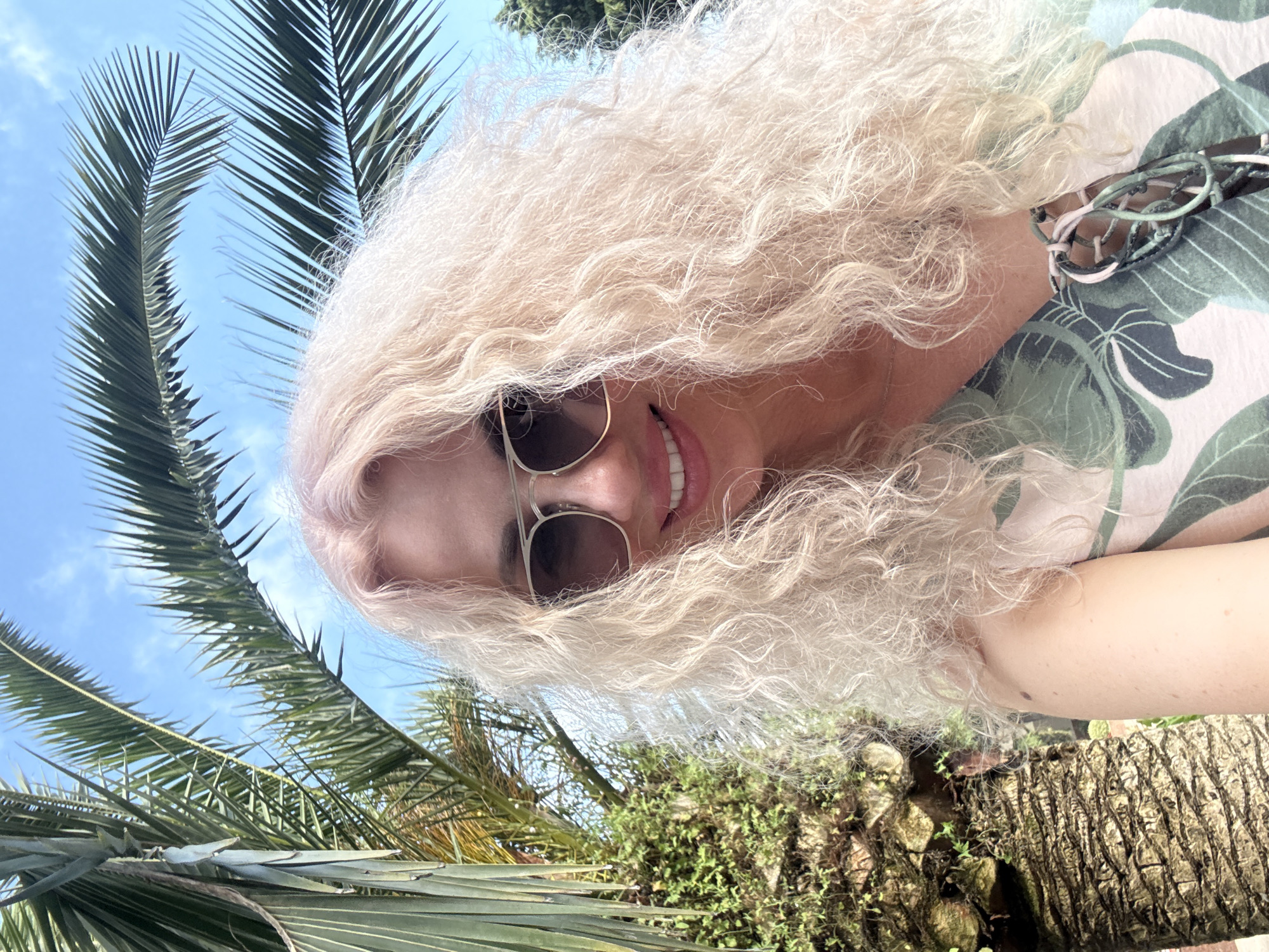 A person with long, curly blonde hair is taking a selfie outdoors. They are wearing sunglasses and a light-colored top with a green leaf pattern. The person is smiling and their teeth are visible. The background shows palm fronds against a bright blue sky with a few clouds, suggesting a sunny day. In the foreground, a palm tree trunk is visible. The overall scene evokes a tropical or subtropical atmosphere, consistent with a location like Sochi, Russia, during daylight hours.