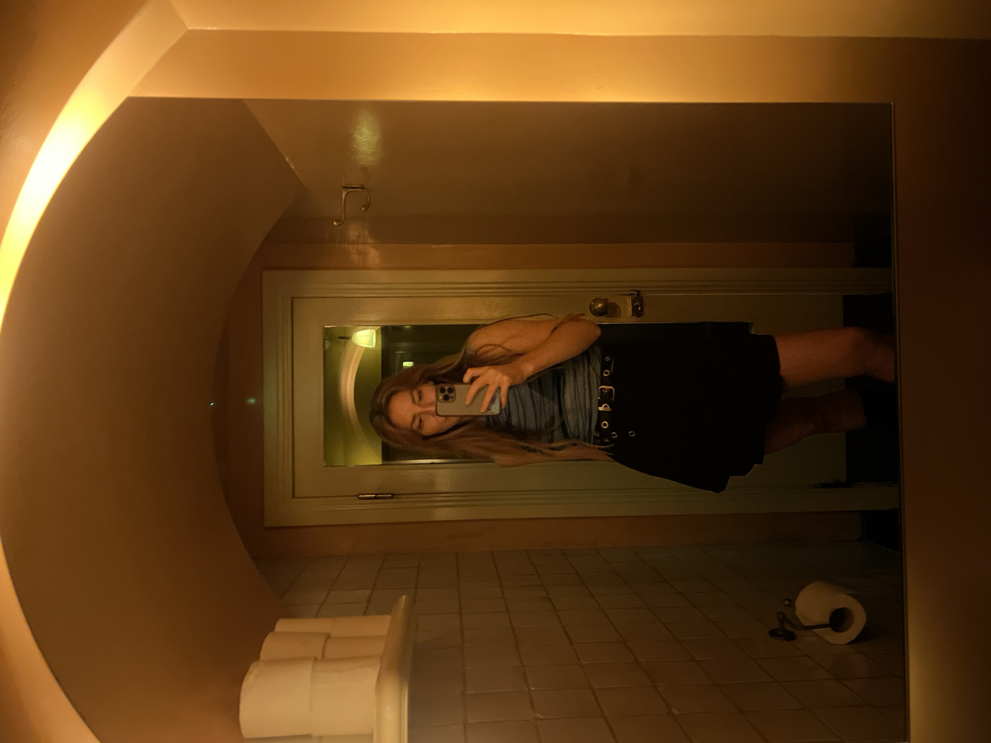 A woman is taking a mirror selfie in what appears to be a restroom. She is wearing a sleeveless blue and white striped top and a black skirt with a belt. Her long brown hair is down. The restroom has tiled walls and a toilet paper dispenser with a roll of toilet paper. There is also a shelf with several rolls of toilet paper on the left side of the image. The lighting is warm and yellow, casting a golden hue over the scene. The background shows a door and more of the restroom's architecture. The location is not specified.