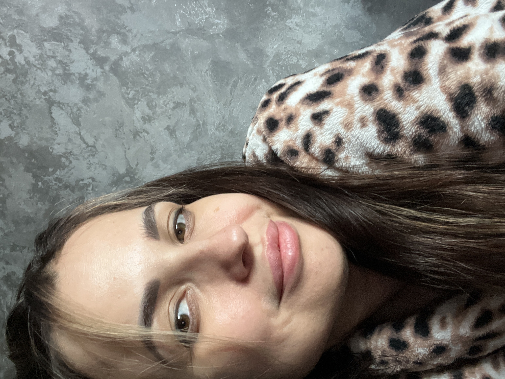 A close-up portrait shows a young adult woman resting or posing against a textured grey wall, partially covered by a leopard print garment. The image is oriented diagonally, with her face filling the central and lower-left portion of the frame. She has long, dark brown hair with lighter highlights, and appears to be looking directly at the camera with her brown eyes. Her eyebrows are well-defined, and she has full, pink lips with a subtle, closed-mouth smile. Her skin is fair, and a minor blemish or scratch is visible on her left cheek. The top of her head and the left side of her face are cropped out of the frame. She is wearing or wrapped in a soft, fuzzy garment featuring a brown and black leopard print pattern against a light beige background, which is prominent in the upper and lower right sections of the image. The background wall has a mottled grey texture, suggesting a decorative concrete or plaster finish. No text is visible.