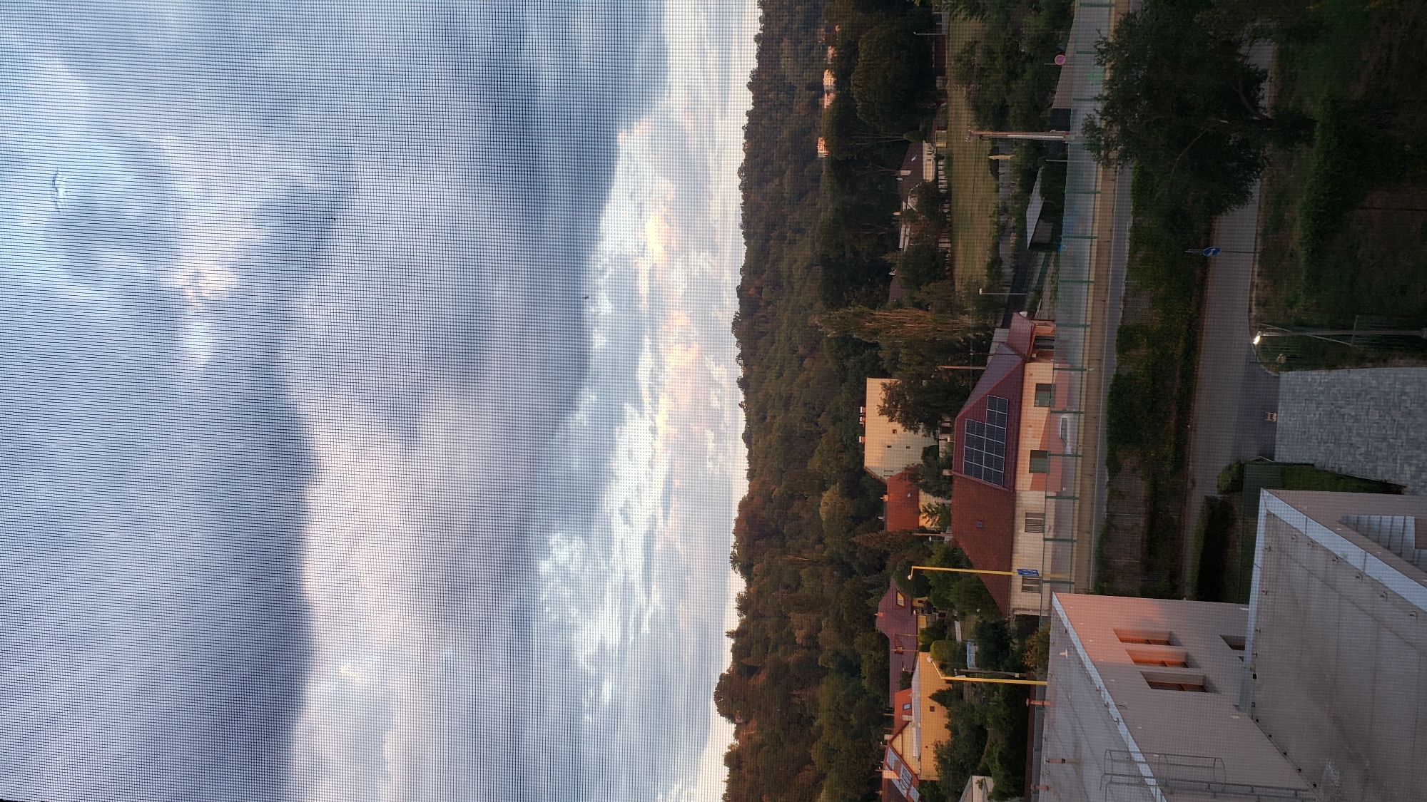 This image depicts a suburban landscape at dusk, viewed through a window screen. In the foreground, the top of a modern building with light-colored walls and multiple windows is visible. Adjacent to it, another building with a flat roof and a walkway leading away from it is partially shown. A gray paved pathway with a lighting fixture is also present.
Further into the scene, a residential area with several houses featuring red-tiled roofs can be seen. One house is notable for its solar panels installed on the roof. Trees and lush green foliage surround these homes, leading up to a densely wooded hillside that dominates the background. A road with a fence runs through the midground, with a white vehicle visible on it.
The sky is filled with dramatic clouds, with streaks of light suggesting either sunset or sunrise. The overall impression is one of a peaceful, residential area on the outskirts of a city. Based on the architectural style and the presence of specific elements common in Slovakian suburban areas, this scene is likely located in the city of Prešov, Slovakia.