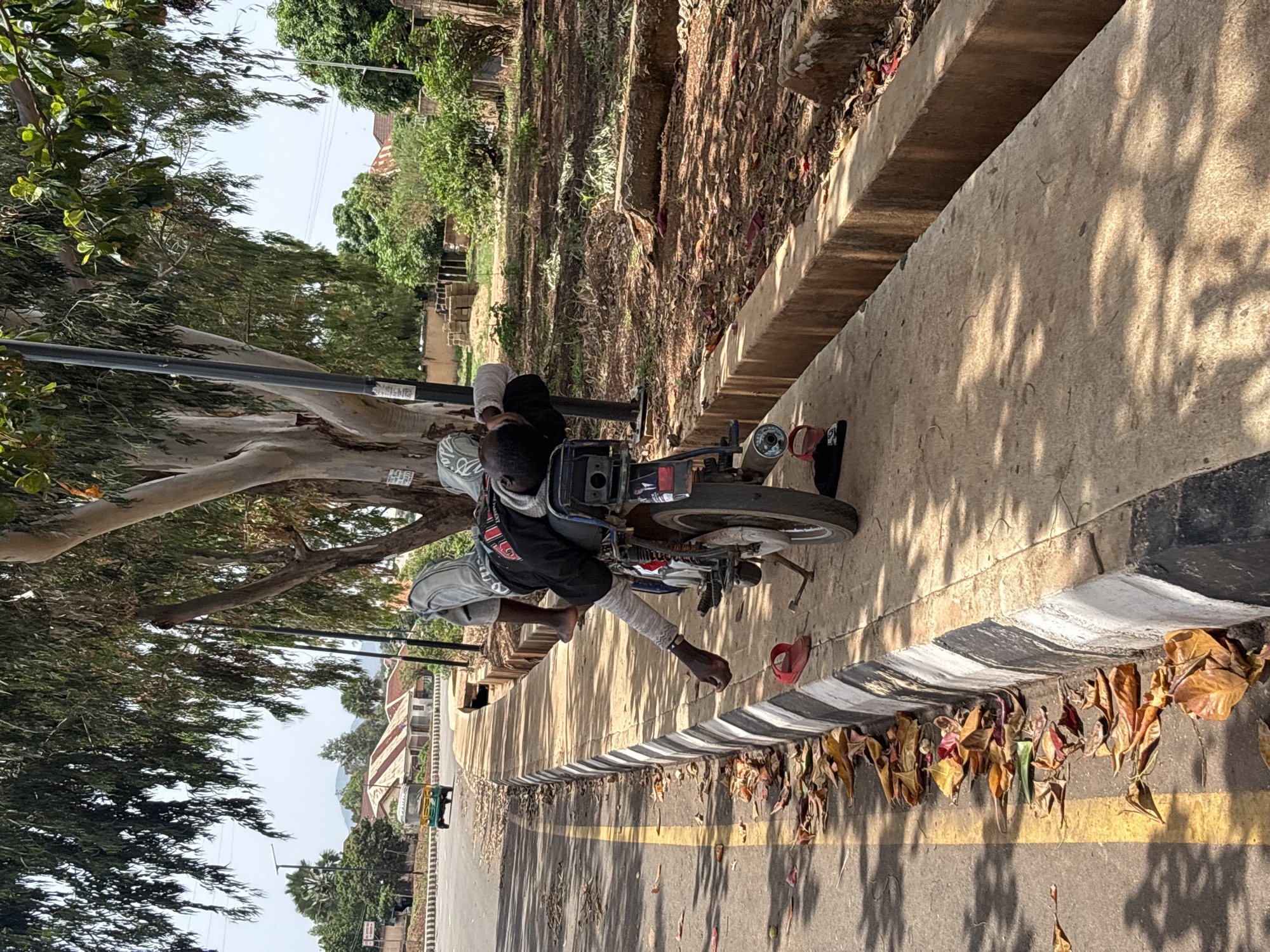 A man sits on a motorcycle parked on the shoulder of a road in Jos, Nigeria. He is wearing a gray shirt, black shorts, and has a bandage on his lower leg. He has bare feet, with a red sandal lying nearby. The motorcycle is parked next to a curb with alternating black and white stripes. Dry leaves are accumulated along the curb. The road has a yellow line marking the shoulder. Trees are visible in the background, and buildings are seen in the distance. A utility pole is situated near the motorcycle. The surrounding environment seems typical for a roadside scene in Jos.