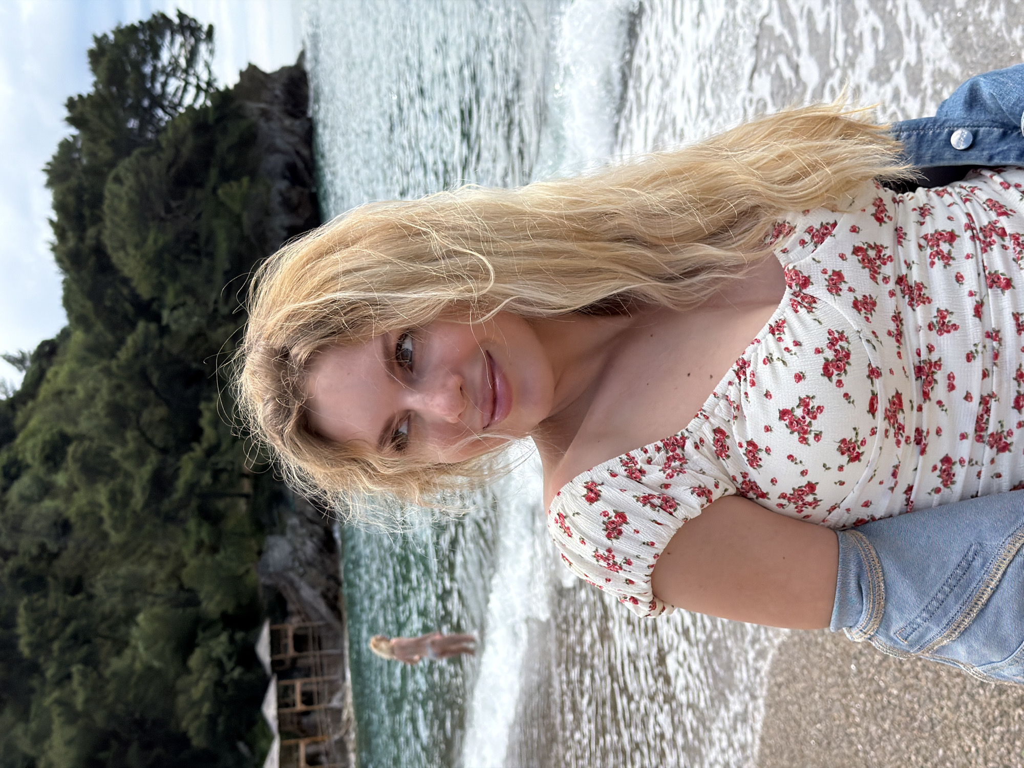 A young woman stands on a beach in Petrovac centar, Montenegro. The woman is blonde and is wearing a floral top and jean jacket. The beach is sandy and the water is clear. In the background, there is a rocky cliff covered in trees. Another person is visible in the water in the background.