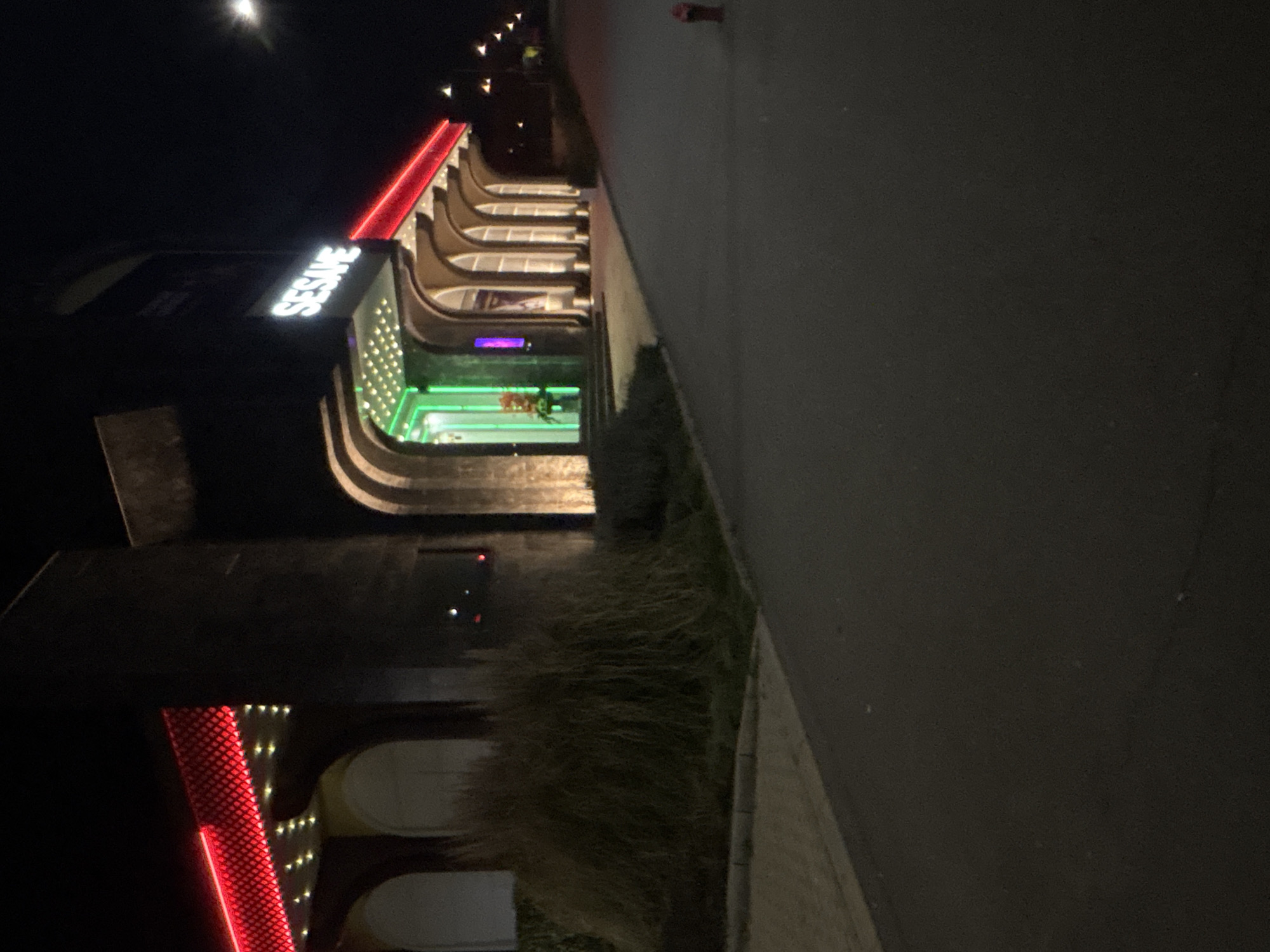 At night, a view of the "Sesame" casino building in Burgas, Bulgaria is captured. The structure features illuminated green lighting and a red stripe across the top, accompanied by white lettering. The casino is adjacent to a paved walkway. Vegetation grows in front of the building and beside the walkway. The street and surrounding darkness surround the building.