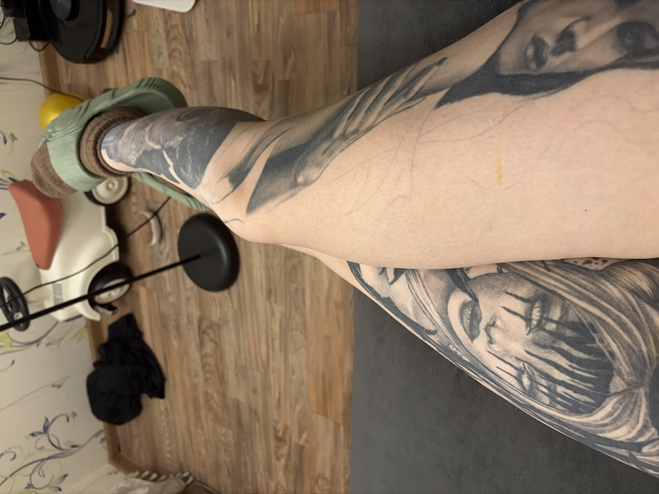 Crossed legs dominate the frame, featuring prominent grayscale tattoos of faces on the skin. The background presents a wooden floor, a child's toy car, a black shirt on the floor, a vacuum cleaner, and a light-green sock. The lighting is indoor and a bit dim. A gray mat is visible.