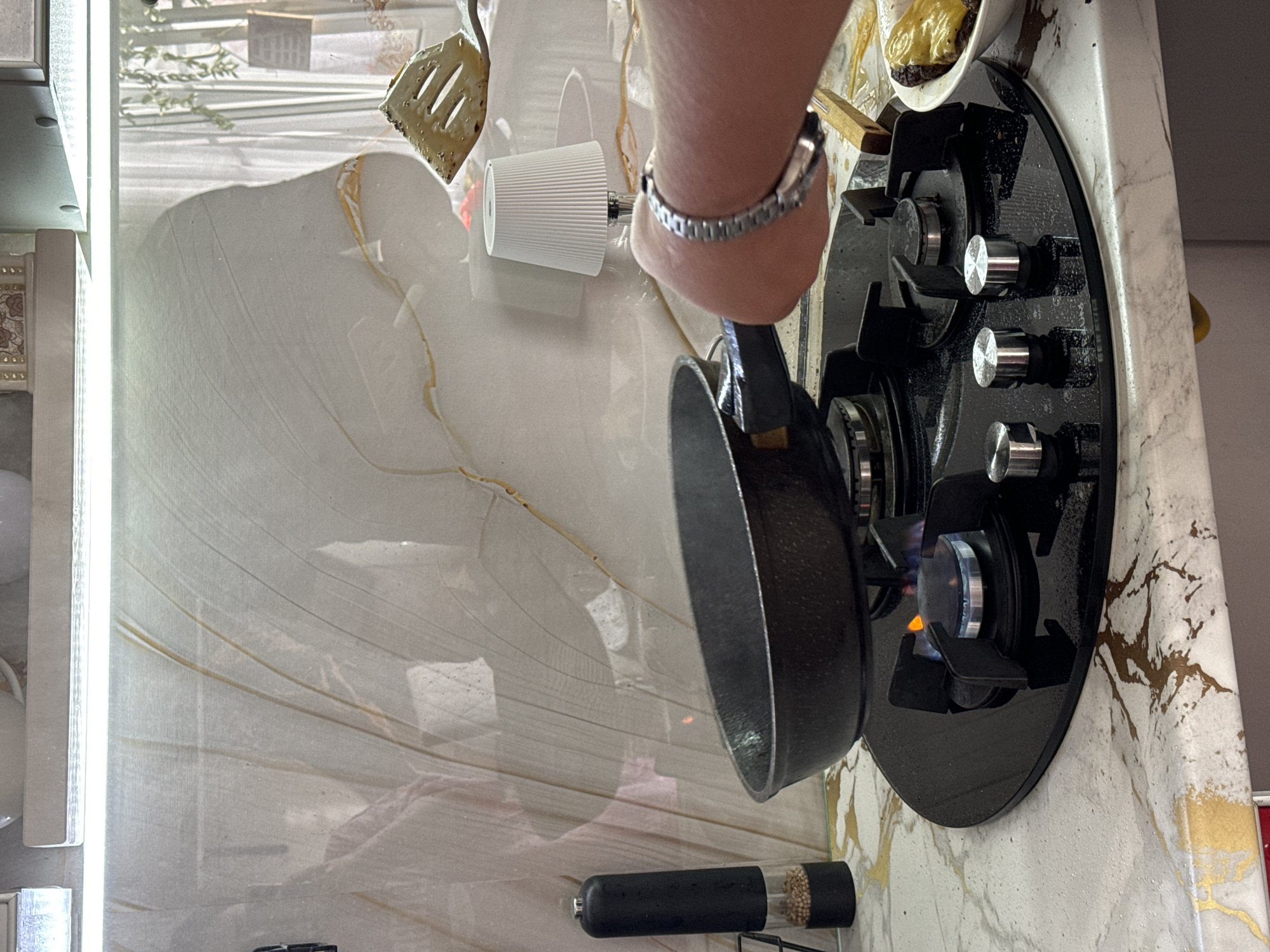 A person's arm and hand are visible, wearing a silver watch, as they hold a small black frying pan over a lit gas burner on a modern black stovetop with silver knobs. The stovetop is set into a marble-patterned countertop with gold veining. To the left of the stovetop, a white ribbed cylindrical object, possibly a salt or pepper grinder, is visible. Further to the left, a white spatula with a wooden handle is resting on the countertop. The background shows a bright, reflective surface with faint patterns, suggesting a kitchen or dining area. The lighting is bright and even, indicating daytime. The overall impression is of food preparation in a contemporary setting.