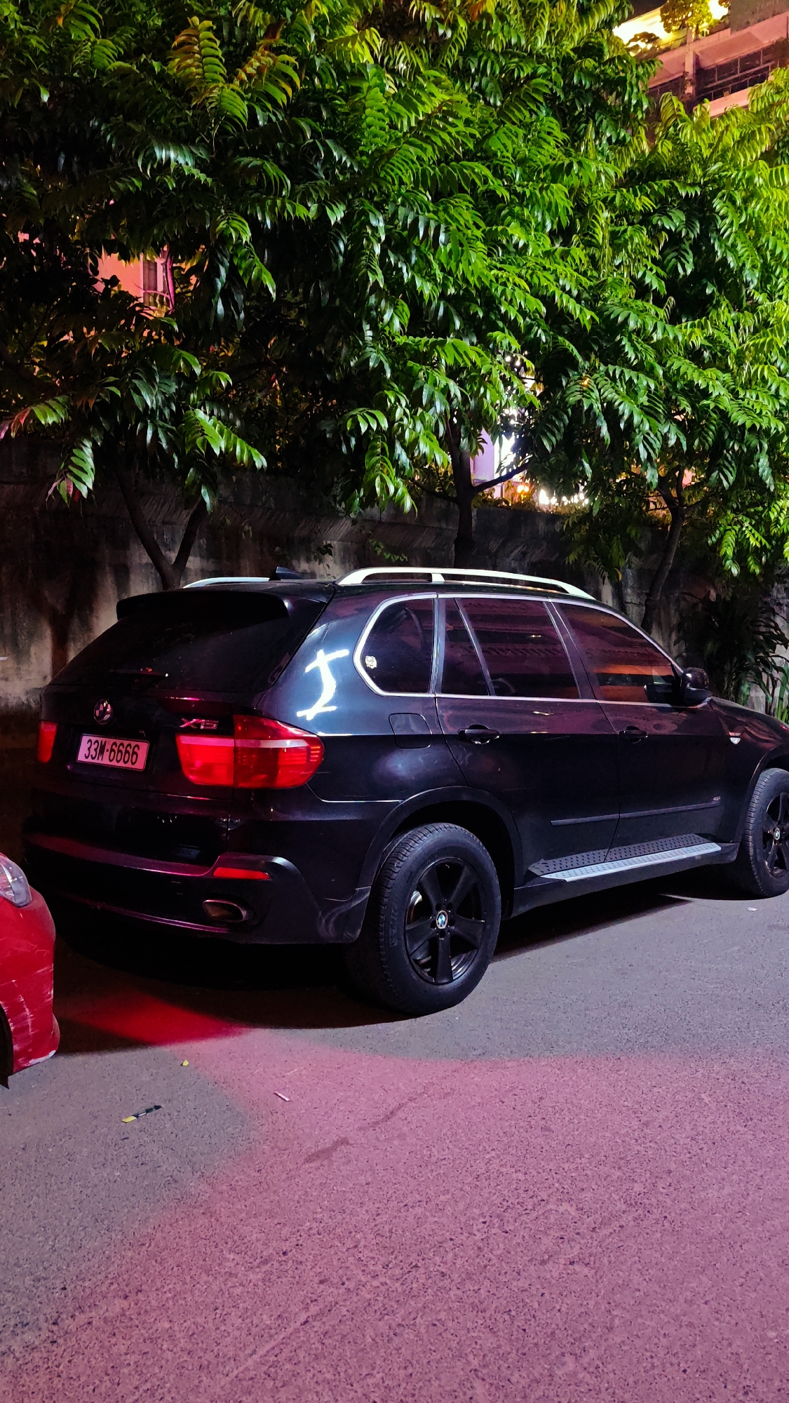 An outdoor, eye-level, medium shot shows the rear right side of a black BMW X5 SUV parked on a street at night. The vehicle's license plate reads "33M-6666". A white stylized marking, possibly a drawing or sticker, is visible on the rear passenger side window. Red light is cast on the ground from the vehicle's taillights and from a red object, likely another vehicle, partially visible on the left. The street is paved and appears to be in an urban setting. Lush green trees with dense foliage are prominent in the background, illuminated from above, suggesting a well-lit area. The overall ambiance is dimly lit with artificial lighting casting vibrant colors on the scene, characteristic of a city like Hanoi, Vietnam, particularly at night when such lighting can be common.