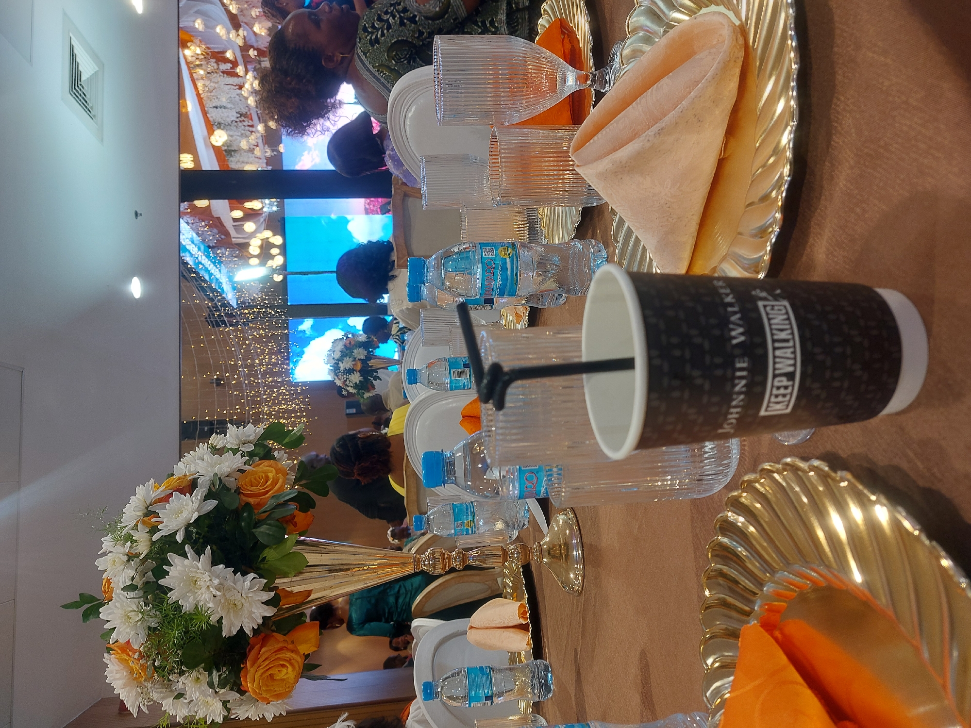 A long banquet table is set with place settings for a formal event. The table features gold-colored charger plates, cloth napkins, water bottles, and various glasses. A tall vase with a floral arrangement of white and orange flowers is a centerpiece. A paper cup with "JOHNNIE WALKER KEEP WALKING" printed on it sits at a place setting. In the background, people are visible, along with a large screen displaying images. The setting appears to be indoors, likely at a banquet hall. Although information is not explicitly available in the picture to confirm, the description asks for a location in Dar es-Salaam, Tanzania. The event may be a wedding reception, gala, or similar formal gathering.