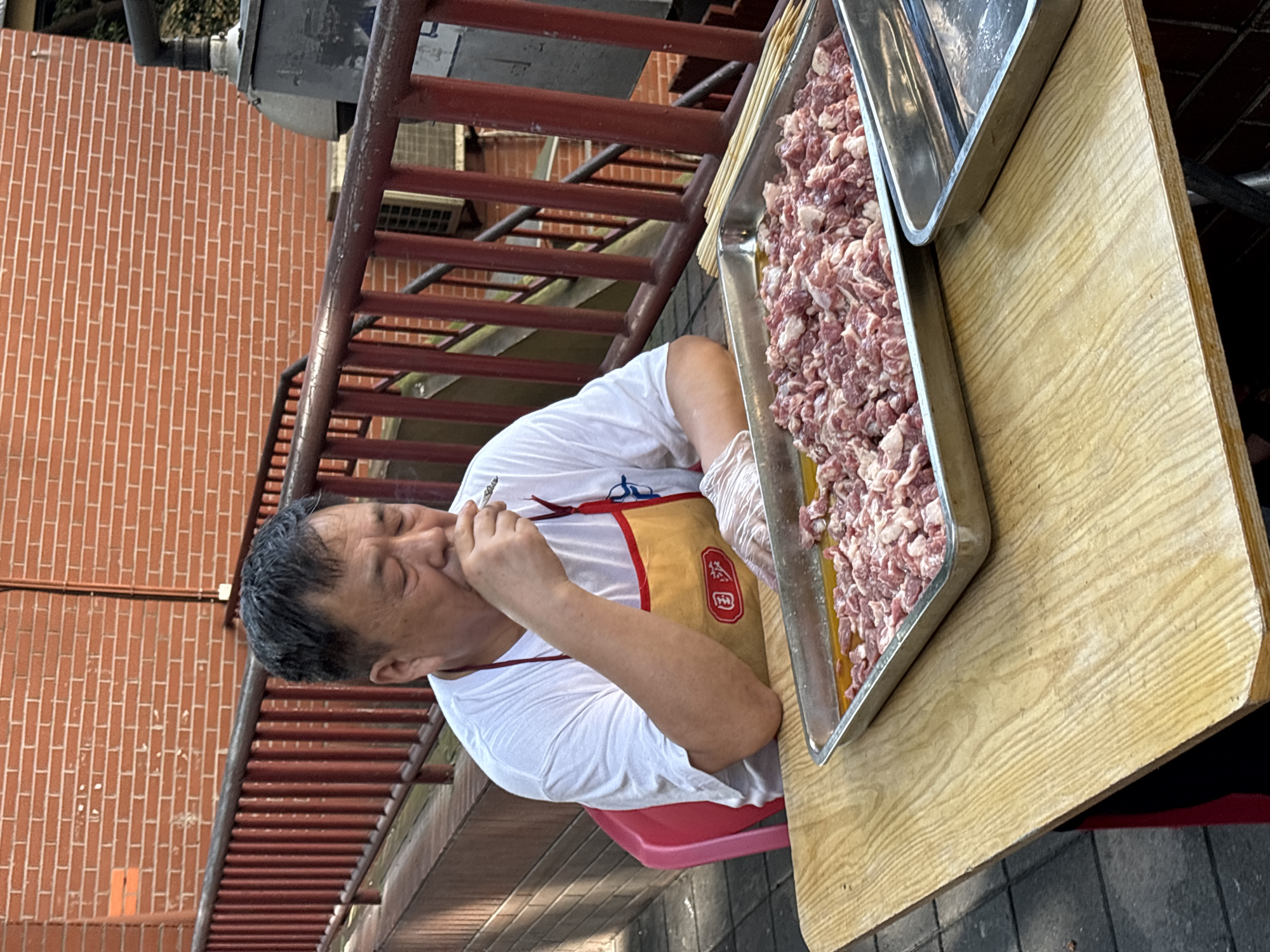 An East Asian man sits at a wooden table outdoors in Yuzhong District, China. He is wearing a white t-shirt and a yellow apron over it. His eyes are closed, and he is holding a cigarette to his mouth, smoking it. He is wearing clear plastic gloves on both hands. In front of him on the table is a large metal tray filled with small, cubed pieces of raw meat. To the right of this tray is an empty metal tray. Behind the man is a red metal railing and a brick wall. A pink plastic chair is to his left. The ground appears to be paved with square tiles.