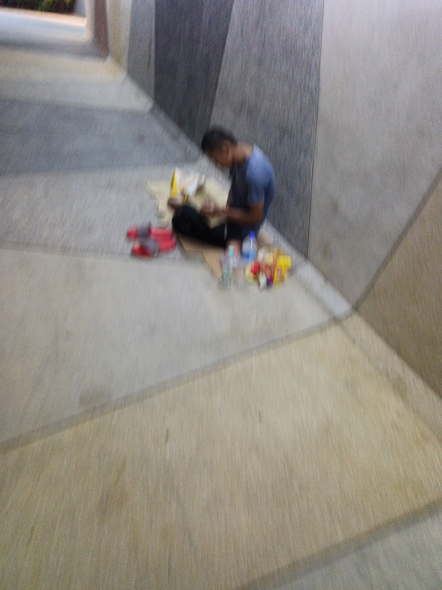 A person is sitting on the ground in a hallway, hunched over and appearing to be reading or working on something. The person is wearing a blue t-shirt and dark pants. In front of the person are several items, including a yellow bag, what appears to be food items, and bottles of water. The hallway has a textured floor and walls with varying shades of gray and beige. The image appears to have been taken at night or in low light, and the blurriness suggests motion. It is not possible to determine the specific location in Kuala Lumpur, Malaysia from the provided image.