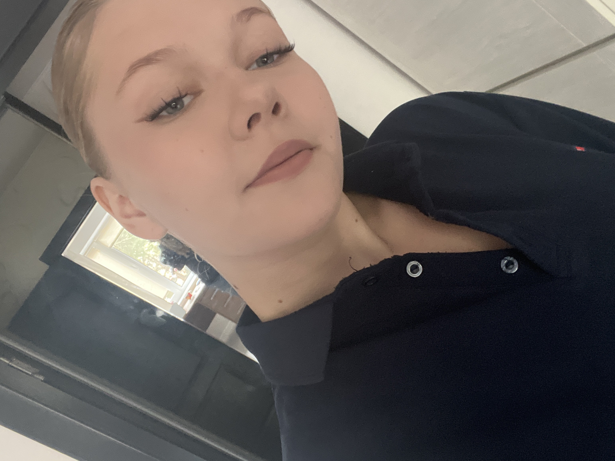 The image depicts a young woman taking a selfie in a dimly lit room. She has blonde hair pulled back and is wearing a dark blue polo shirt. Her makeup includes winged eyeliner and neutral lipstick. The background shows a dark door on the left, a mirror reflecting a window with daylight, and a sliver of a wall on the right. The overall impression is a casual, indoor setting, possibly a bathroom or dressing area. There are no discernible elements that specifically indicate the location is Perm, Russia.
