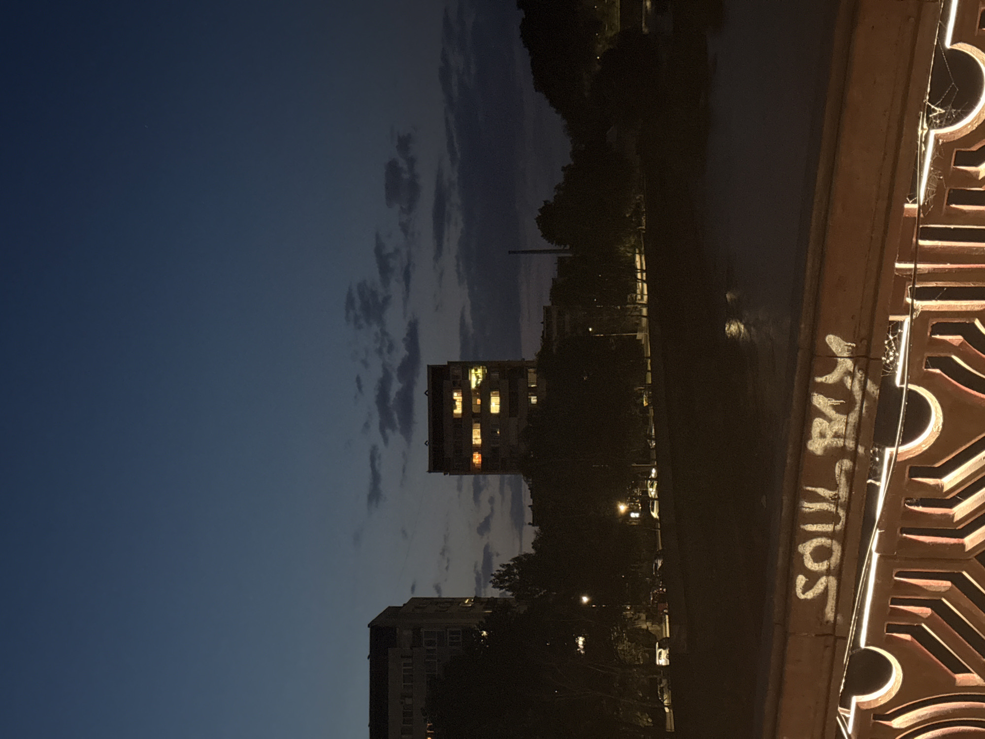 This is a nighttime photograph of an urban landscape, likely taken from a bridge or elevated vantage point, overlooking a body of water. In the middle ground, two apartment buildings are visible, one taller and to the left, the other slightly shorter and to the right. Both buildings have lit windows, suggesting occupancy. Trees and other vegetation form a dark band between the buildings and the water. In the foreground, a railing is visible, adorned with graffiti that reads "SOUL RYM". The sky is a deep blue, with scattered clouds, indicating dusk or twilight. The scene appears to be in the city of Lovech, Bulgaria, possibly near the Osam River, given the presence of water and the characteristic architecture of Eastern European apartment blocks. There are no people clearly visible in the image. The overall impression is one of a quiet, urban evening.