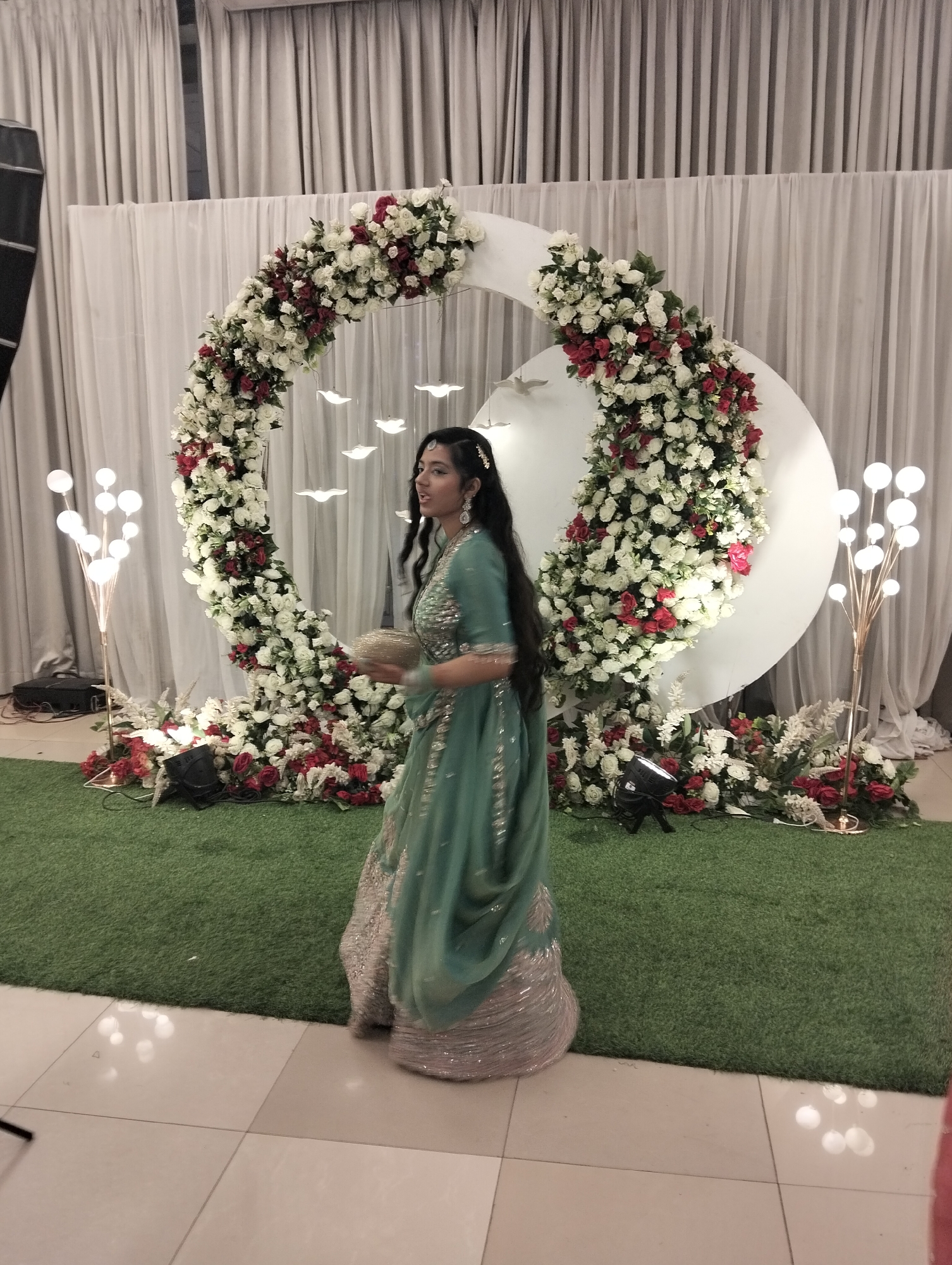 The image shows a woman in a teal, silver, and gold dress, facing away and holding a gold container in her hands. She appears to be standing in front of a circular floral arrangement of red and white flowers. The arrangement is set against a white backdrop in front of white drapes. There is also a small patch of artificial grass. On either side of the floral arrangement are light fixtures with multiple bulbs. The location appears to be indoors and is in the city of Unknown, Unknown.