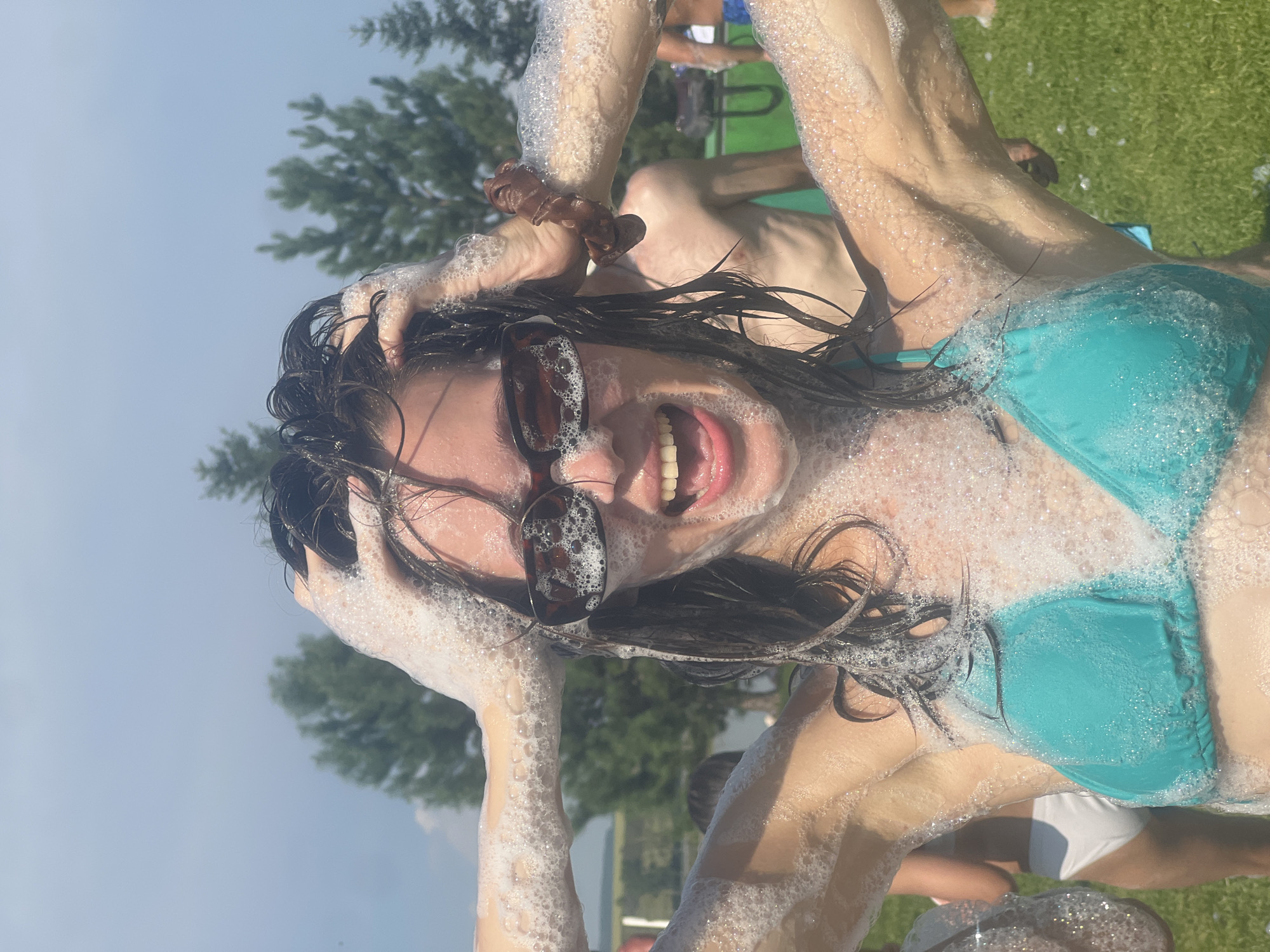 A young woman with dark hair, wearing sunglasses and a turquoise bikini top, is pictured from the chest up outdoors. She is laughing with her mouth open and has her hands on her head, which is covered in soap suds. Bubbles are also present on her arms and face. In the background, other people can be seen, some of whom are also covered in suds. The environment appears to be a grassy area with trees and a body of water, possibly a lake, visible behind the subjects. The sky is cloudy. The scene suggests a recreational activity, likely a foam party, taking place in a natural setting.