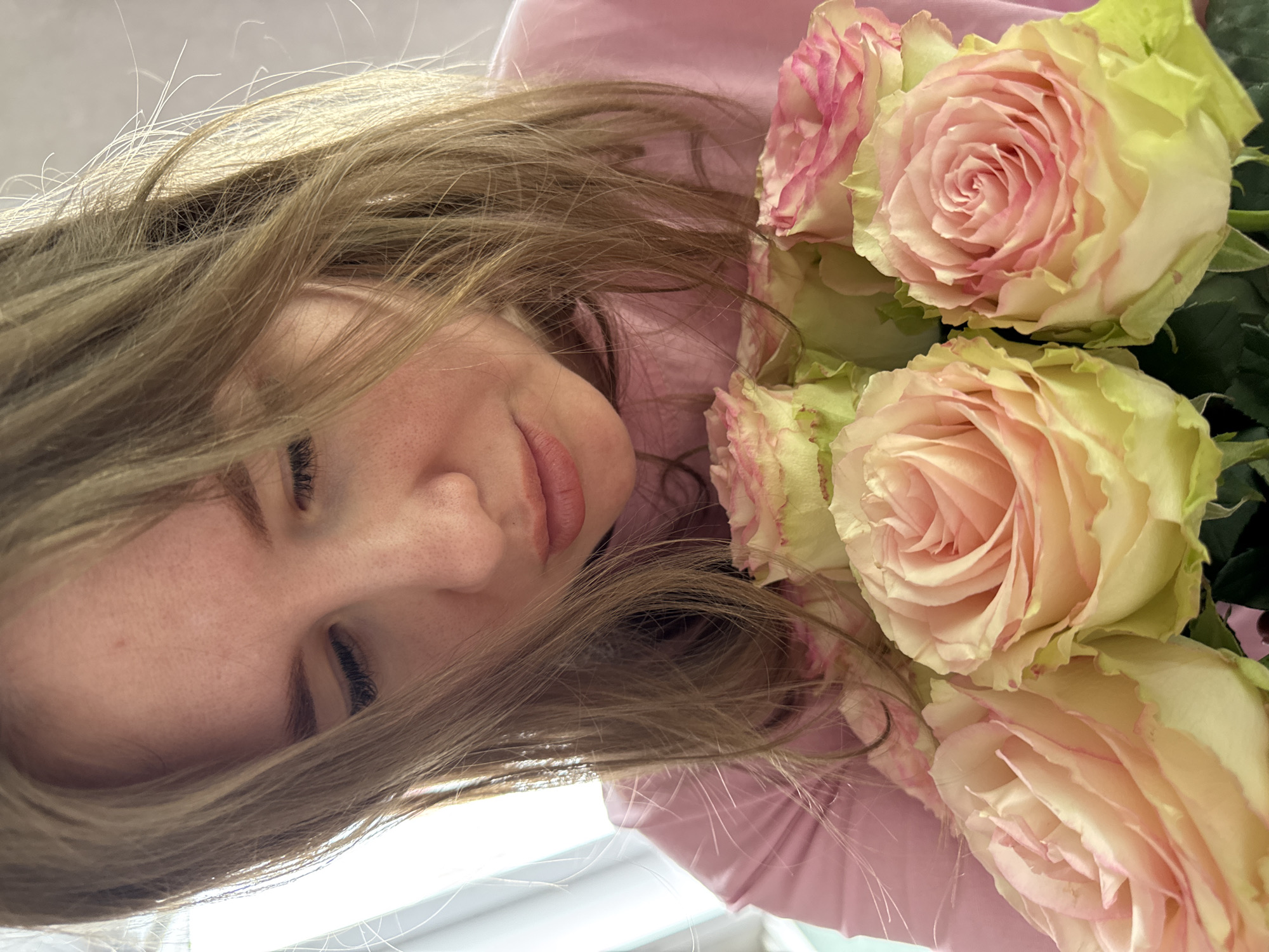 The image depicts a close-up, head-and-shoulders view of an adult female holding a bouquet of roses indoors in Gornyi (Dvurechenskoe S/P), Russia. The female, positioned off-center to the right, has light brown hair cascading over her shoulders and partially obscuring her face. Her head is tilted downwards, and her eyes appear closed or heavily lidded, directing her gaze towards the flowers. She is wearing a pink long-sleeved top. In the lower foreground, a cluster of approximately six light pink and cream-colored roses with pale green outer petals is prominently displayed. The background is indistinct, brightly lit by an unseen light source to the left, suggesting a window or bright interior space. No explicit interactions are visible with other individuals.