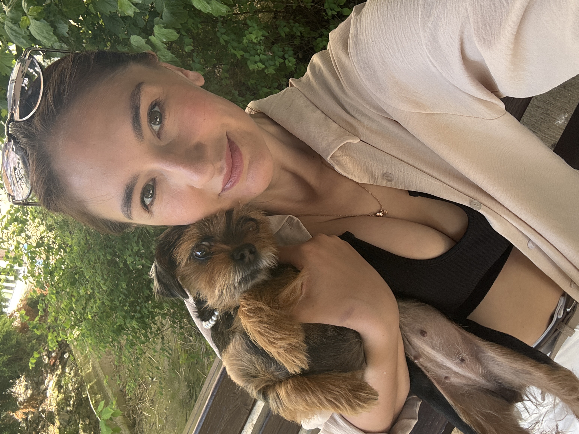 A woman is holding a dog in a selfie. The woman has sunglasses perched on her head, light brown hair, and is wearing a tan collared shirt over a black top. She is smiling. The dog is a brown and black terrier mix. The background shows foliage, suggesting an outdoor setting, possibly a park or garden. Based on the prompt, this is likely Sochi, Russia.