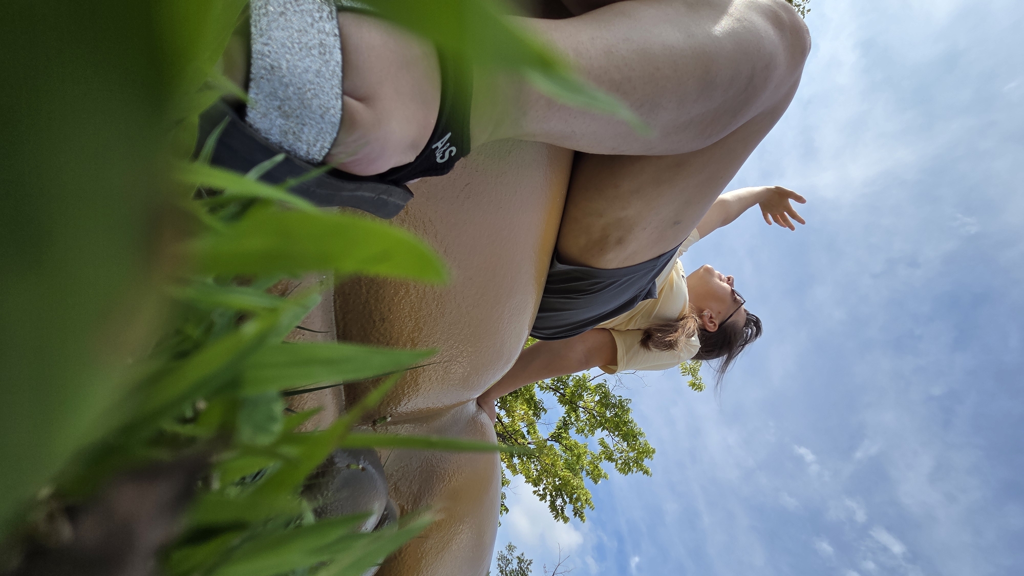 A person is lying on a large, golden, anatomical sculpture in Dunnae-myeon, South Korea, with their legs bent and arms raised towards the sky. The person is wearing a yellow t-shirt and dark shorts, and sunglasses. They appear to be looking up at the sky with their hand shielding their eyes from the sun. The sculpture is positioned outdoors, in a grassy area with trees in the background. The perspective is from a low angle, with blades of grass in the foreground obscuring part of the sculpture and the person's feet. The sky is blue with scattered white clouds.