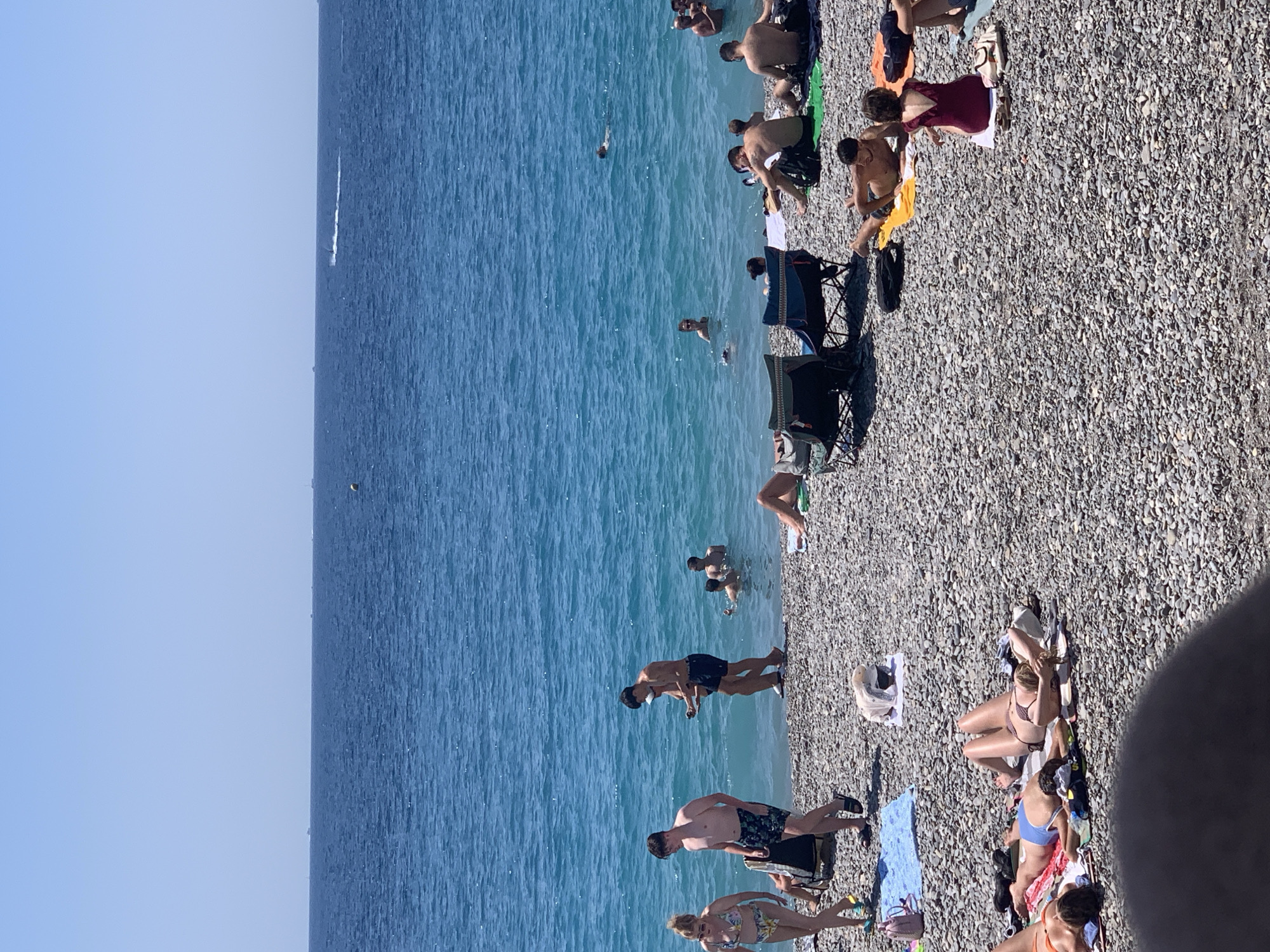 This is a ground-level, eye-level, medium shot photograph taken on a sunny day at the pebble beach of Nice, France, along the Mediterranean Sea. The water is a vibrant turquoise blue, with small waves lapping against the shore. The beach itself is covered in grey pebbles.
In the foreground, several people are sunbathing on towels and chairs. Some are lying down, others are sitting. A woman in a red swimsuit is sitting on a white towel, facing away from the camera. To her left, a man in black swim trunks is lying on a yellow towel.
Further back, closer to the water, more people are visible. A group of men are standing in the shallow water, some are talking. A few individuals are swimming or wading in the sea. On the beach, two people are walking towards the water. A woman in a bikini walks on the left, and a man in black swim trunks walks behind her.
In the middle ground, a man in a blue shirt and black shorts is standing near the water's edge, looking down. A man in a white cap and black shorts is beside him. Several beach chairs are set up on the pebbles.
In the background, the vast expanse of the blue sea stretches out to the horizon, under a clear blue sky. A few distant boats or buoys are visible on the water.
The overall scene depicts a lively summer day at a popular beach location.