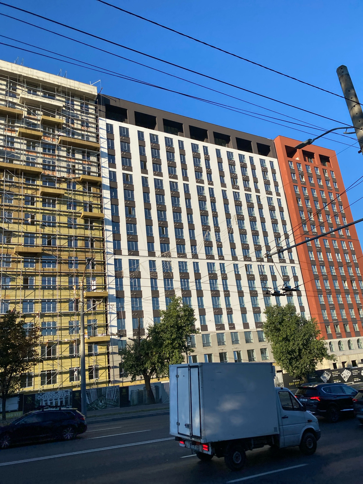 The image shows a view of a modern apartment building complex under construction and partially completed in Chișinău, Moldova. On the left, a section of the building is covered in scaffolding and insulation, indicating ongoing construction. Adjacent to it, a completed section features a white facade with dark-trimmed windows. Further to the right, another section of the building is clad in burnt orange panels, also with dark-trimmed windows. The building is tall and multi-story, with a clear blue sky in the background. Overhead power lines crisscross the upper portion of the image. In the foreground, a white delivery van is parked or driving on a road, with a dark-colored SUV visible behind it. The scene is set in an urban environment, with trees and streetlights also present. A sign with a phone number is visible on a fence near the construction site.