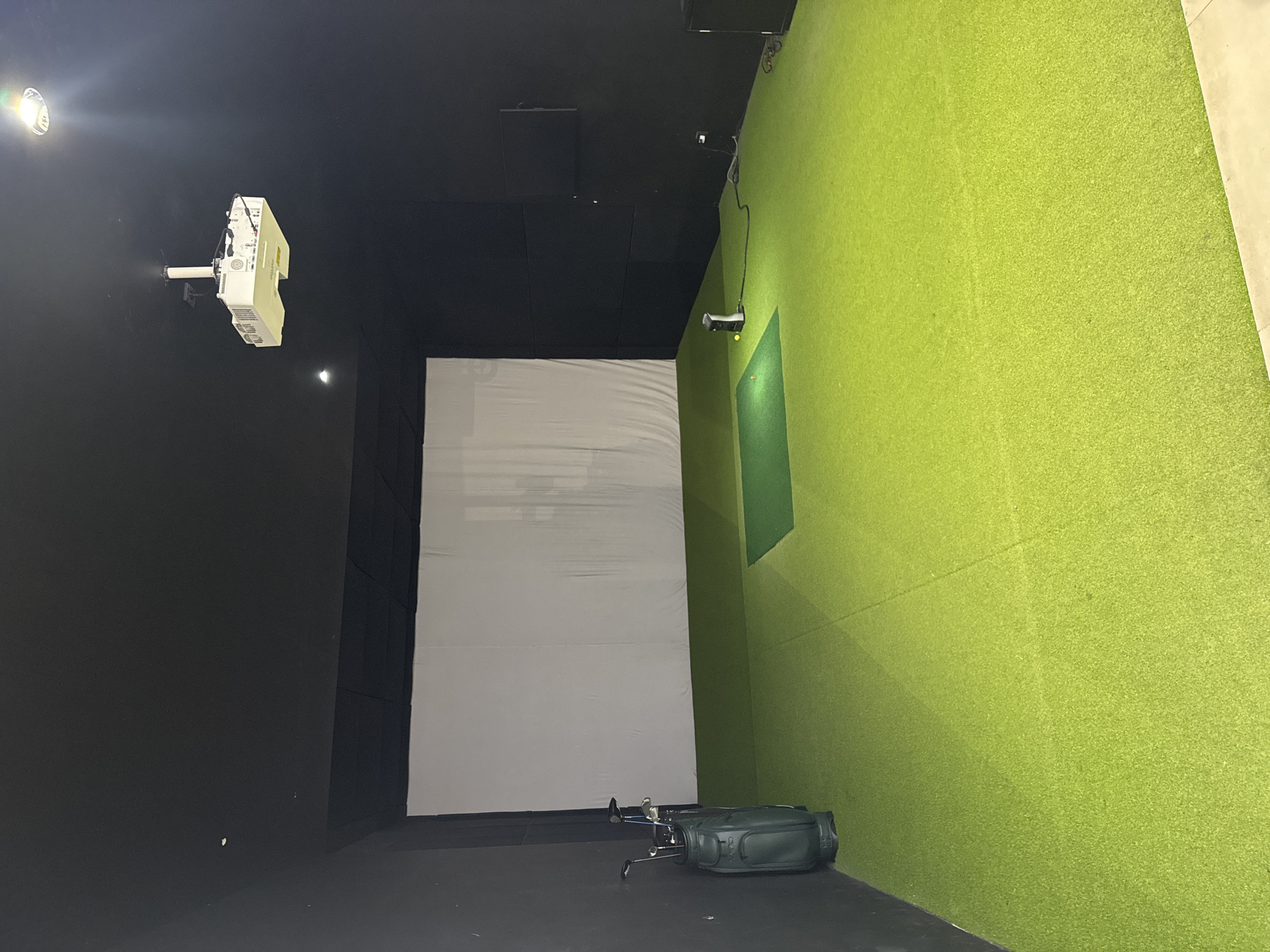 This is an indoor golf simulator room. The room has black walls and ceiling, with a white screen at the far end. The floor is covered in bright green artificial turf. On the left side of the room, a dark green golf bag stands filled with golf clubs. In the center of the room, a small green mat is placed on the turf, with a golf ball and tee on it. To the right of the mat, a projector is mounted on the ceiling, and a white device with a black cord is on the floor. A television screen is mounted on the back wall on the right. The scene is brightly lit by overhead lights.