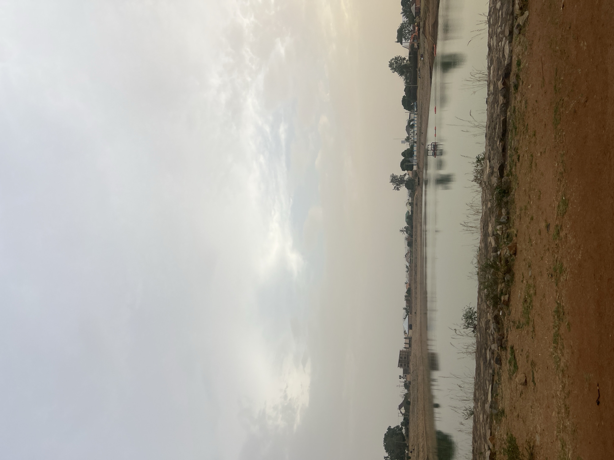 The image shows a body of water in Bukuru, Nigeria, reflecting a hazy, overcast sky. The shoreline consists of a dirt bank interspersed with rocks and sparse vegetation. Buildings and trees are visible in the distance along the opposite bank of the water body. There are no people visible in the image.