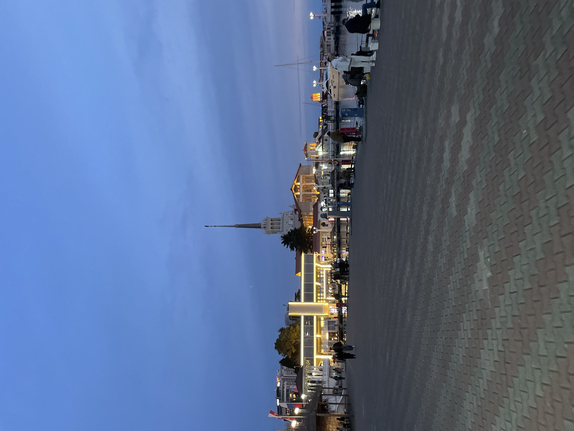 At dusk, the cityscape of Sochi, Russia, is seen from a ground-level perspective on a pedestrian-paved surface. The brickwork is a pattern of gray and red. The sky is a deep blue. Illumination comes from the buildings, which include a prominent steeple. Some people are walking. Along the waterfront, boats are visible.