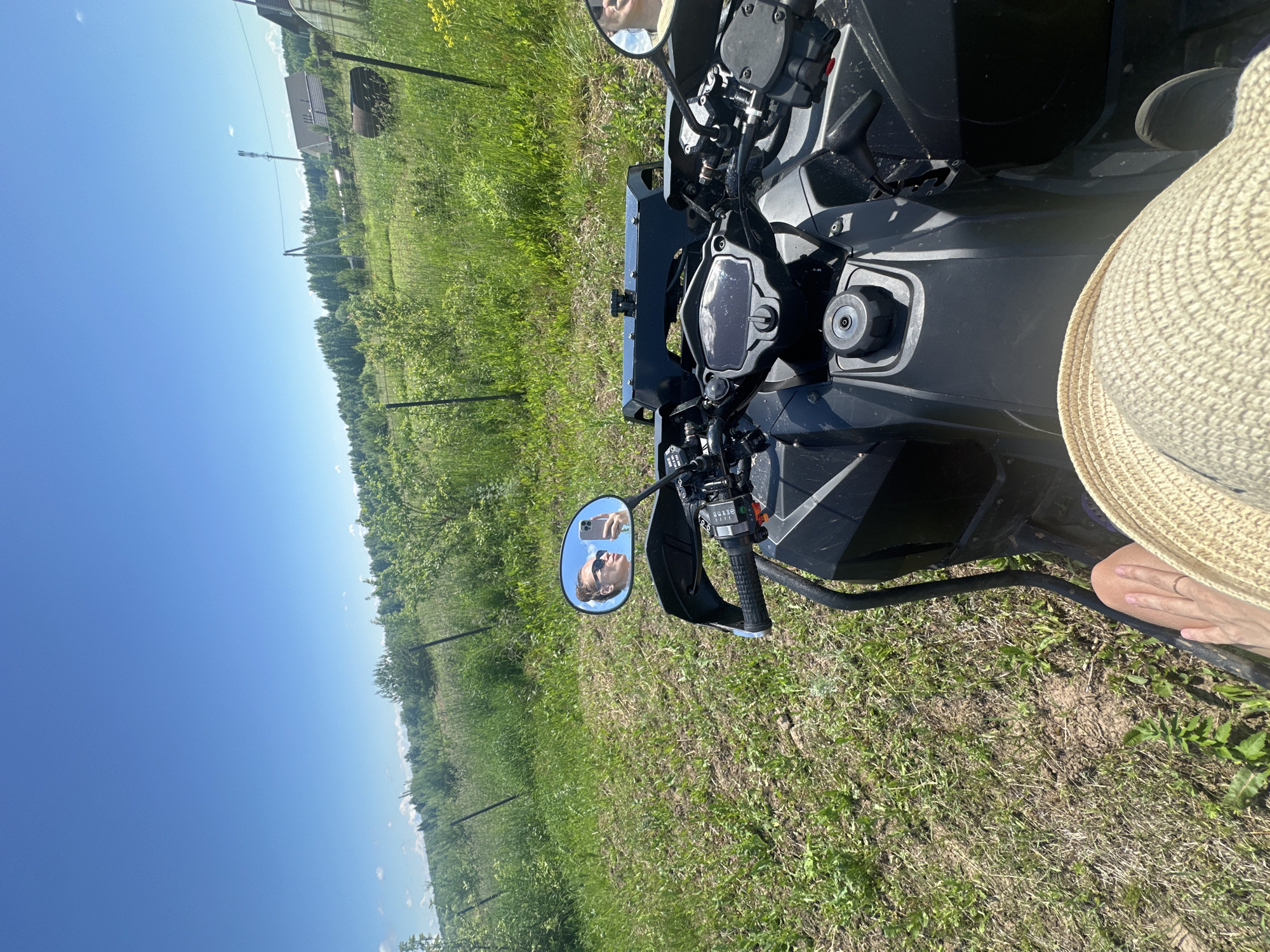 The image captures a first-person perspective of someone sitting on a black ATV, in a rural outdoor environment, likely Kharnavy, Russia. The ATV's handlebars and dashboard are visible in the foreground, along with the rider's straw hat and a ring on their finger. A mirror on the ATV reflects the rider, a woman with her hair in a bun, wearing sunglasses and holding a smartphone, seemingly taking a selfie. The background shows a grassy field with a fence, trees, and some distant buildings under a clear blue sky.