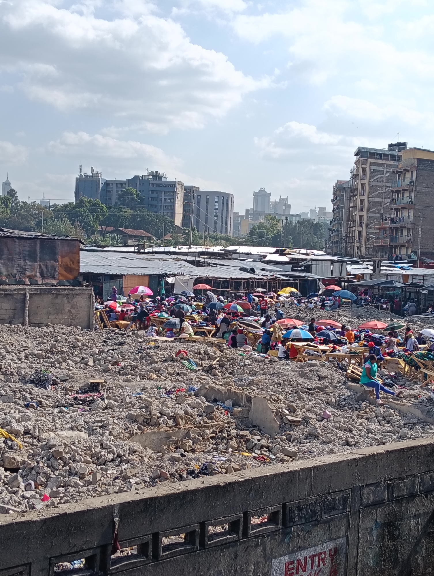 This is a wide shot of a bustling outdoor market in Nairobi, Kenya. The foreground is dominated by a pile of rubble, suggesting recent demolition or construction. In the mid-ground, a vibrant marketplace thrives amidst the debris. Numerous stalls, covered with corrugated iron roofs and fabric awnings, are packed with people. Umbrellas of various colors – pink, red, blue, and yellow – are scattered throughout the market, providing shade for shoppers and vendors. People are seen browsing, selling goods, and interacting. Wooden tables and makeshift stands display clothing and other merchandise. Beyond the market, the cityscape of Nairobi rises with a mix of modern high-rise buildings and older, more weathered structures, indicating the urban setting. The sky is partly cloudy, with sunlight illuminating the scene.